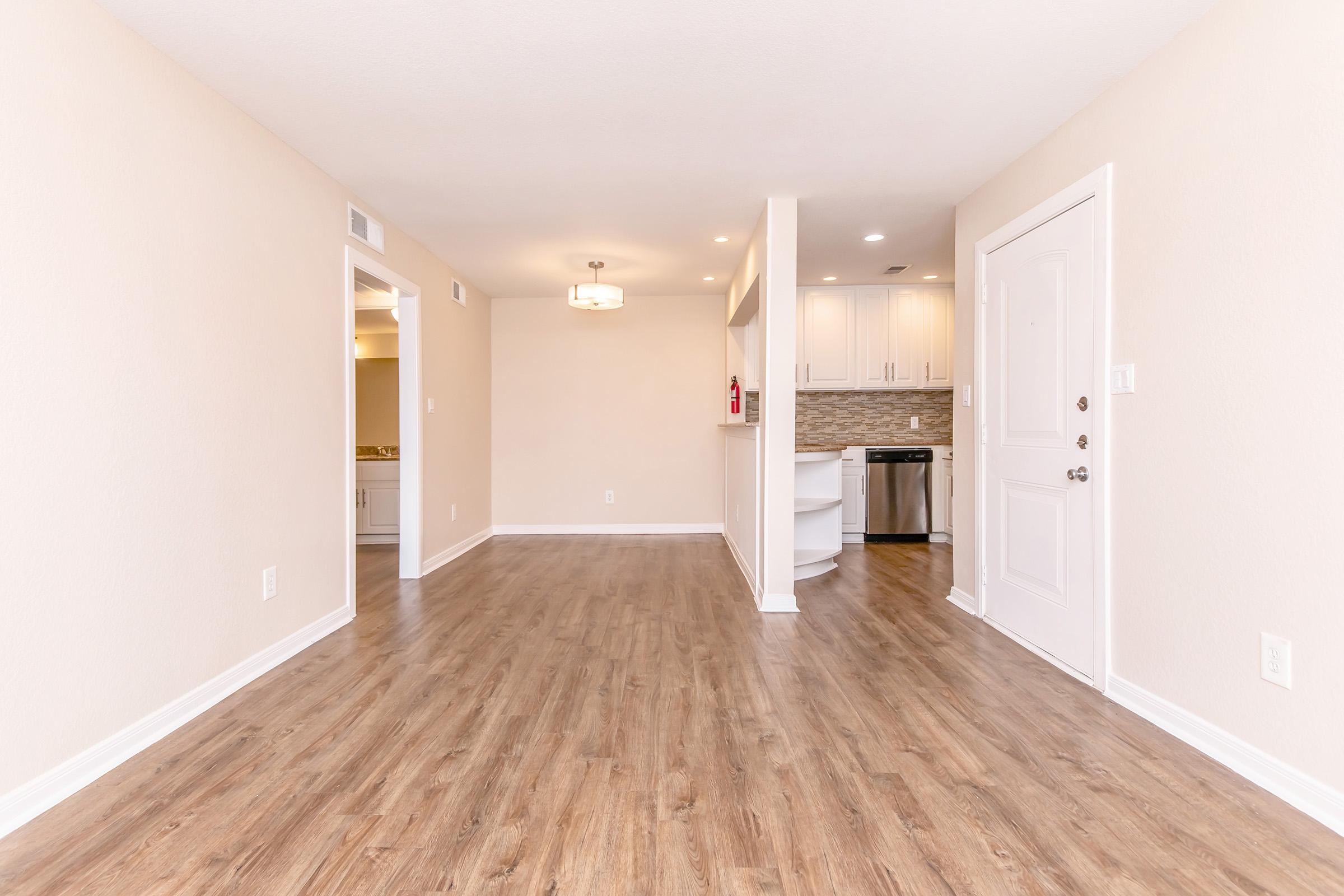 Interior view of a modern, empty living space with light brown wooden flooring. The walls are painted in a soft beige color. A small kitchen area is visible on the right, featuring white cabinets and stainless steel appliances. The entrance door is on the right side, and there is a ceiling light fixture.