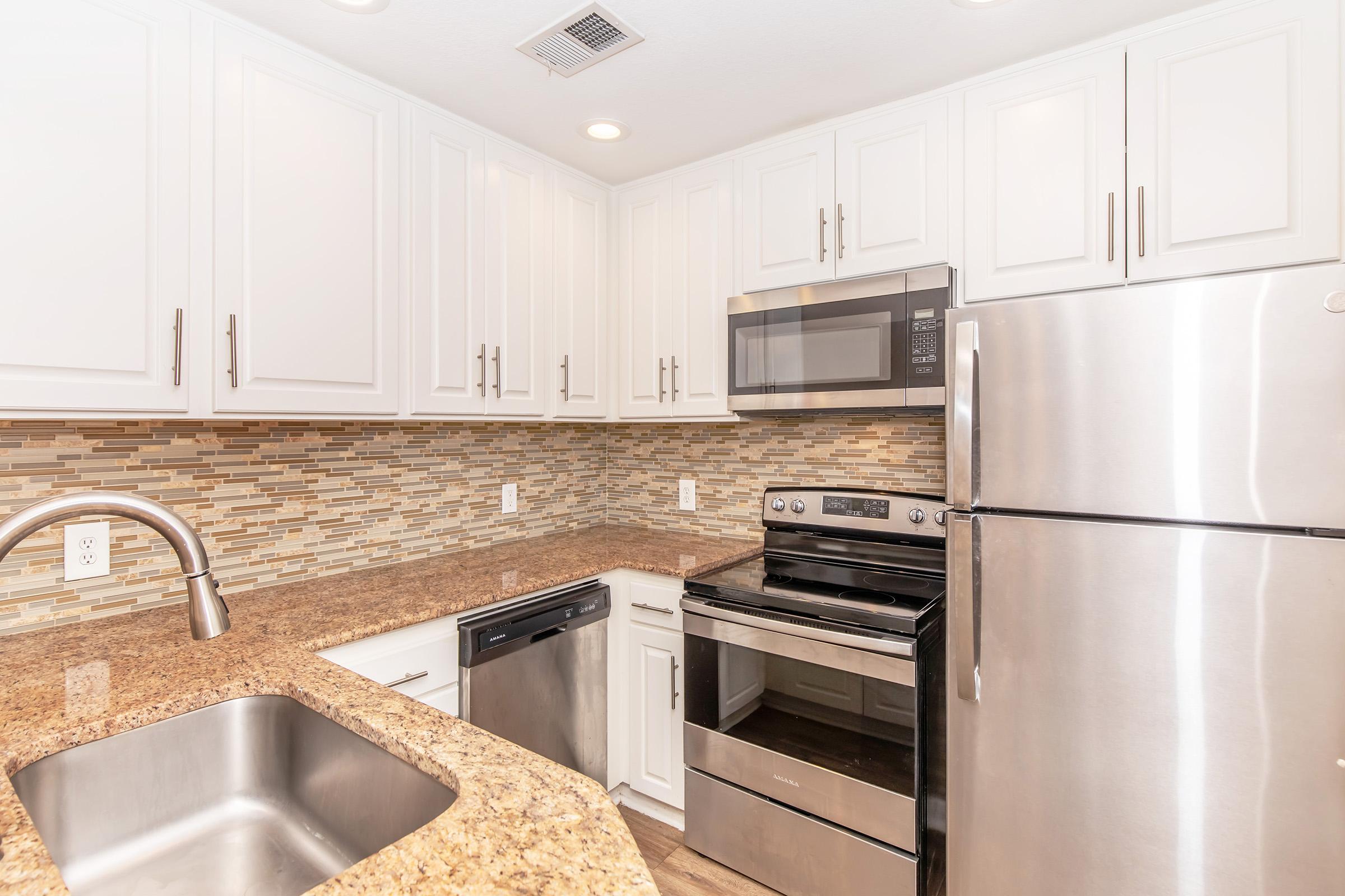 Modern kitchen featuring white cabinets, stainless steel appliances, and a granite countertop. A sink with a faucet is visible, along with a microwave, oven, and refrigerator. The backsplash consists of small, multi-colored tiles, enhancing the contemporary design of the space. Bright lighting adds to the welcoming atmosphere.