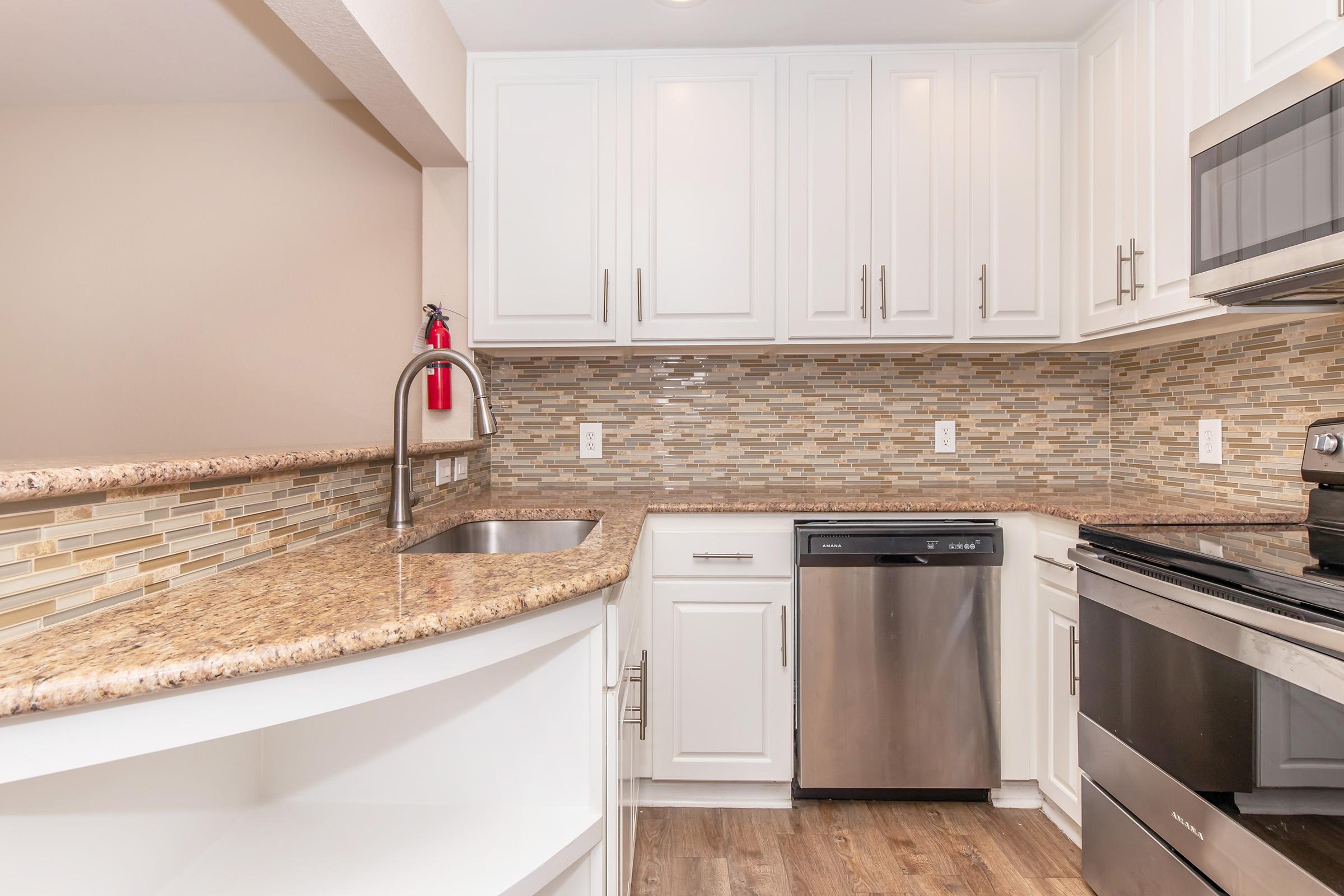 Modern kitchen featuring white cabinetry, a granite countertop, and a stylish backsplash. Stainless steel appliances include a dishwasher and a microwave. The space is well-lit and has an open layout, with a sink integrated into the countertop and a fire extinguisher mounted on the wall.