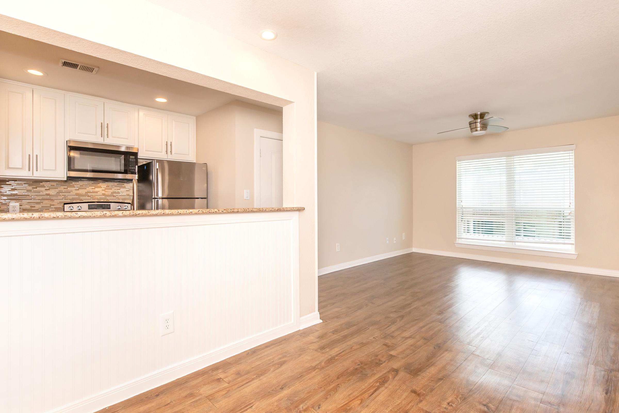A spacious, modern living area featuring an open concept design. The kitchen is equipped with stainless steel appliances and white cabinetry, complemented by a tiled backsplash. The adjacent living space is bright with natural light from large windows and has elegant wood flooring.