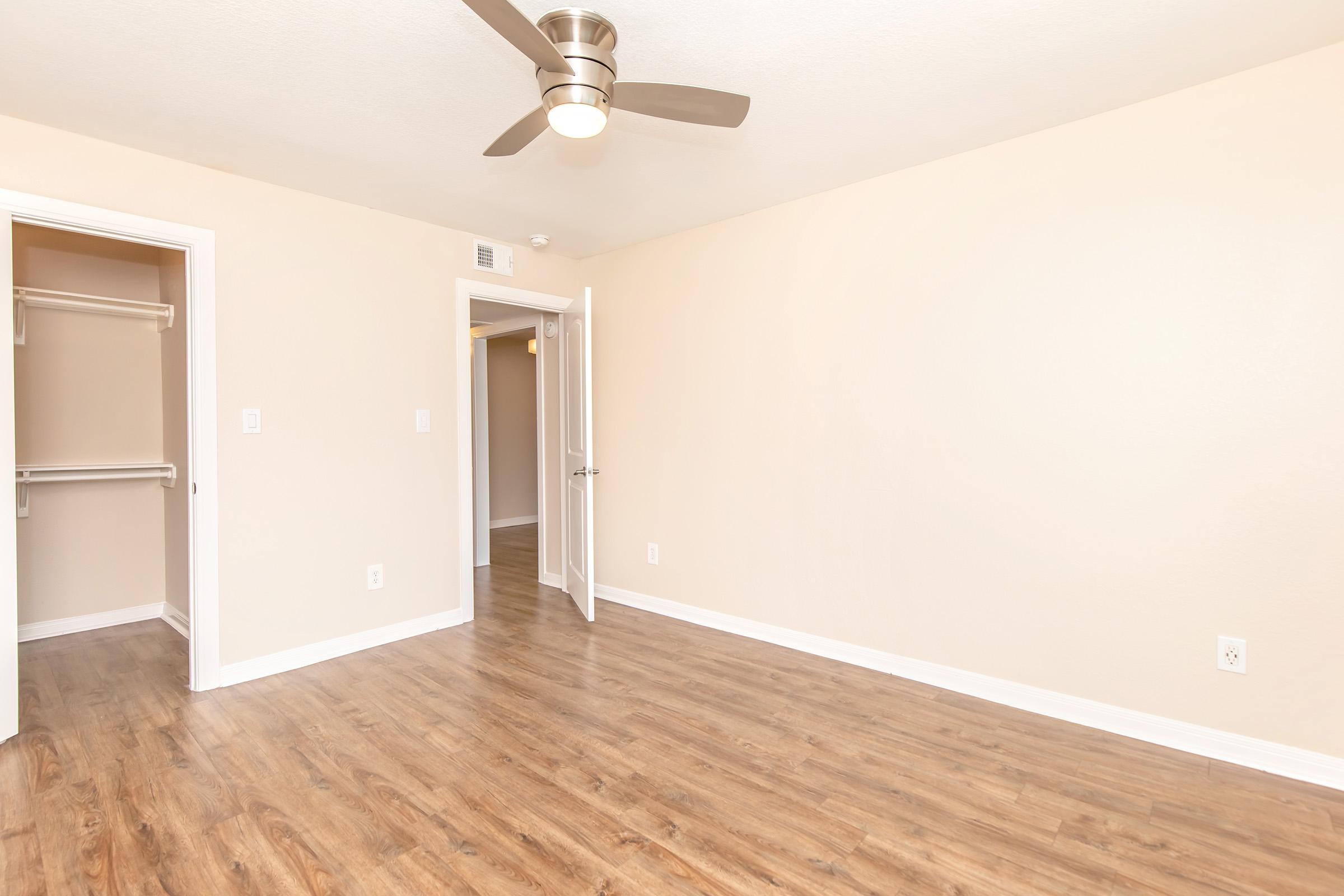 A spacious, empty room with light beige walls and a wooden floor. A ceiling fan is visible, providing a modern touch. An open door leads to another room, and a nearby closet with an open door is partially visible. The overall atmosphere is bright and inviting, ideal for personalizing.