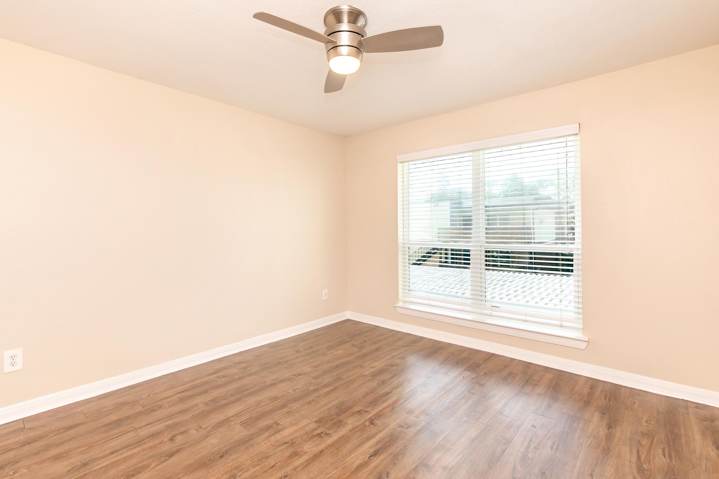Empty room with light beige walls and a smooth wooden floor. A ceiling fan with metal blades is installed, and there is a large window with white blinds, allowing natural light to enter. The window offers a view of an exterior space, enhancing the room's brightness and openness.