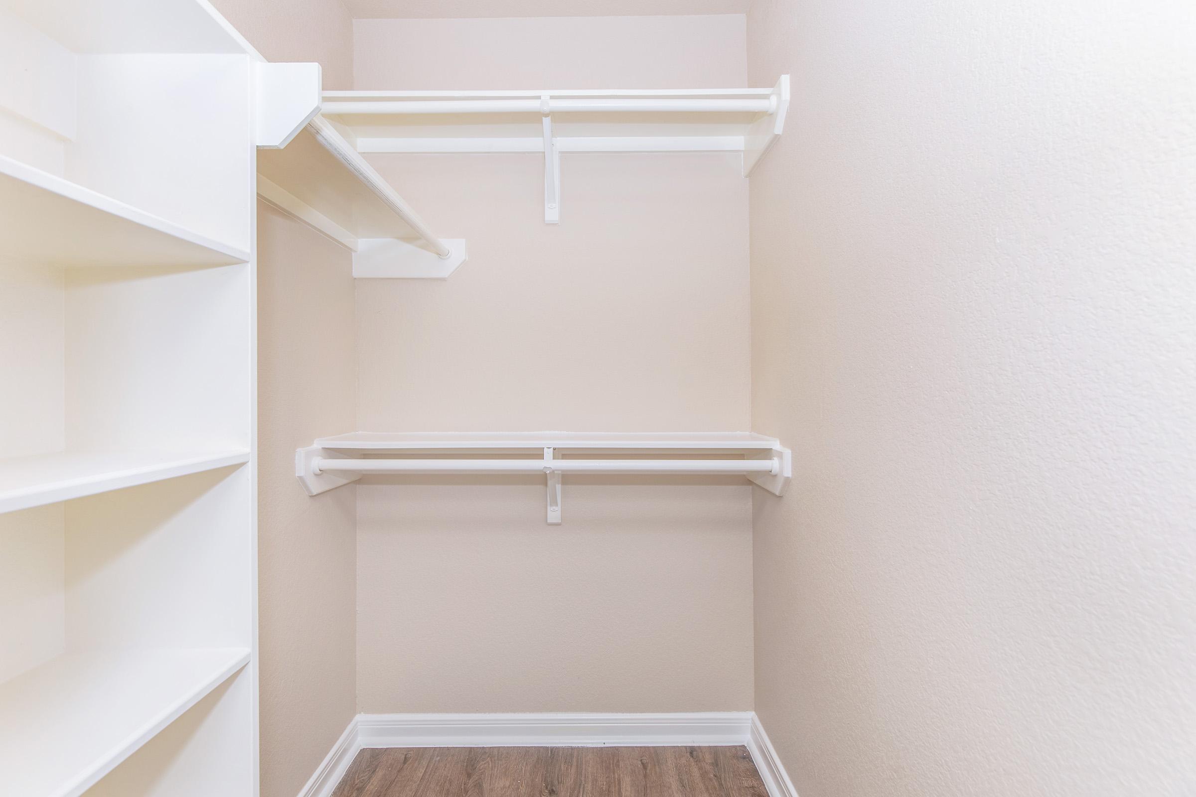 A clean, empty closet with two hanging shelves and one built-in shelf on the left side. The walls are painted in a light beige color, and the floor has a wood-like finish. The space is uncluttered, providing ample storage opportunities.