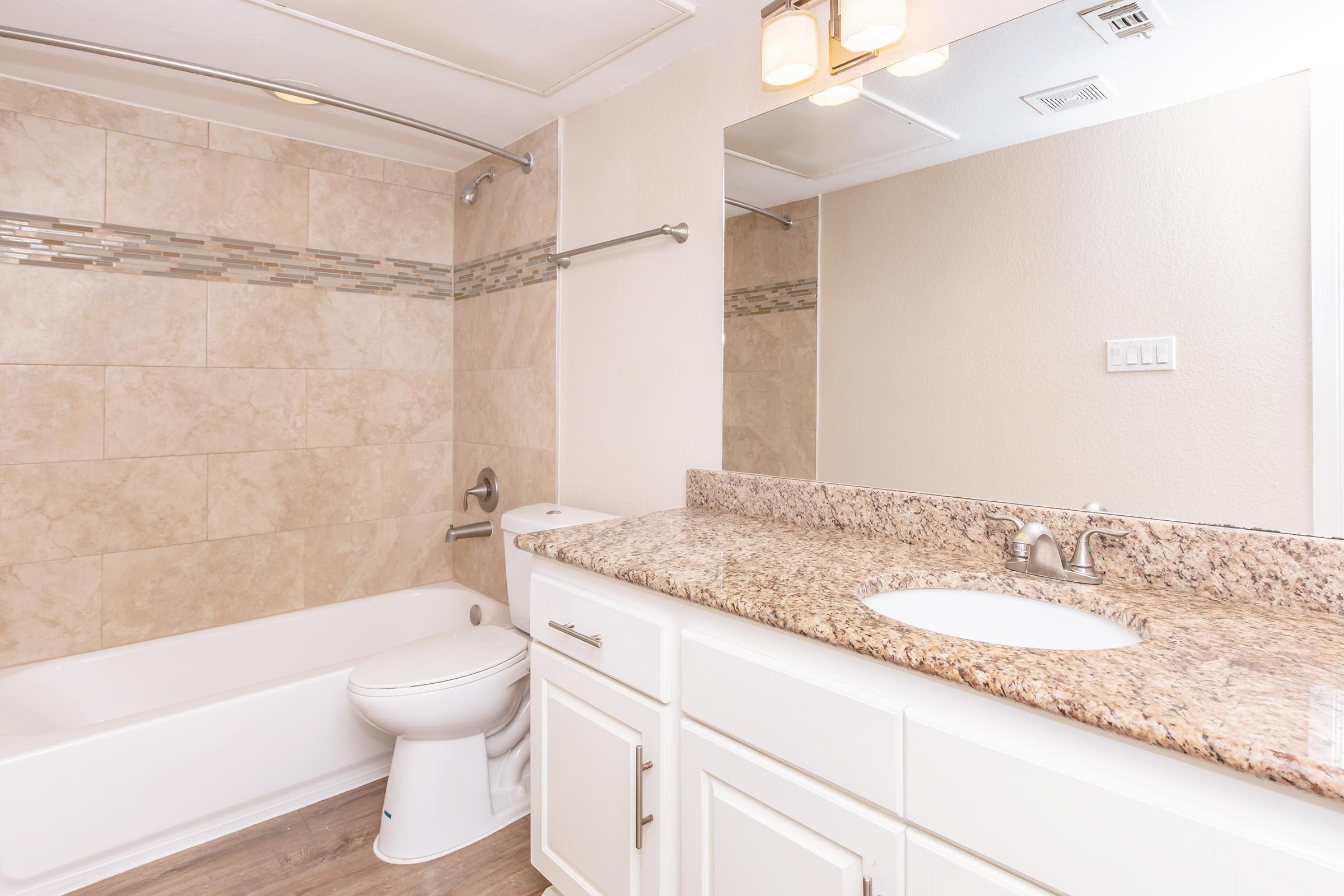 A modern bathroom featuring beige tiled walls, a white bathtub, and a granite countertop with dual sinks. The area has a large mirror above the sink and stylish lighting fixtures. The floor has a wood-like appearance, adding warmth to the space.