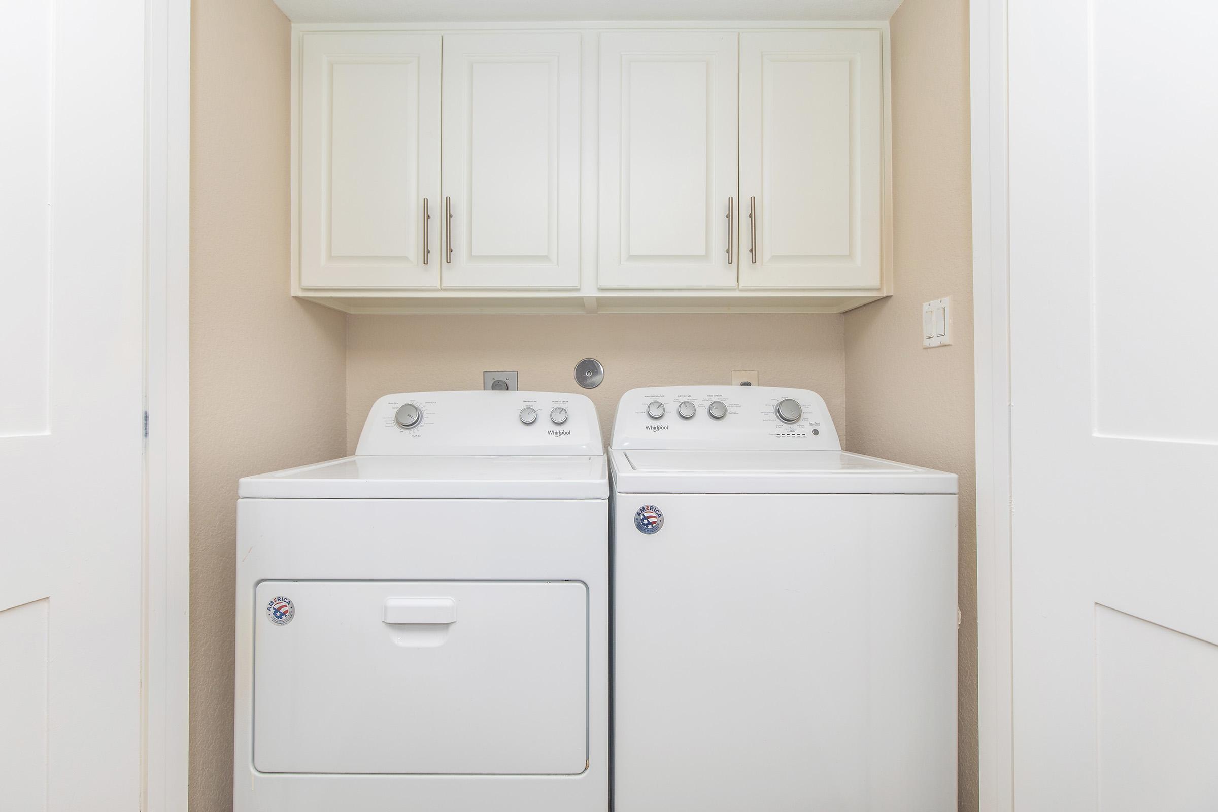 A laundry room featuring a stacked white washer and dryer set against a light beige wall. Above the appliances, there are white cabinets with minimalist handles for storage. The overall space is clean and well-organized, suitable for laundry tasks.