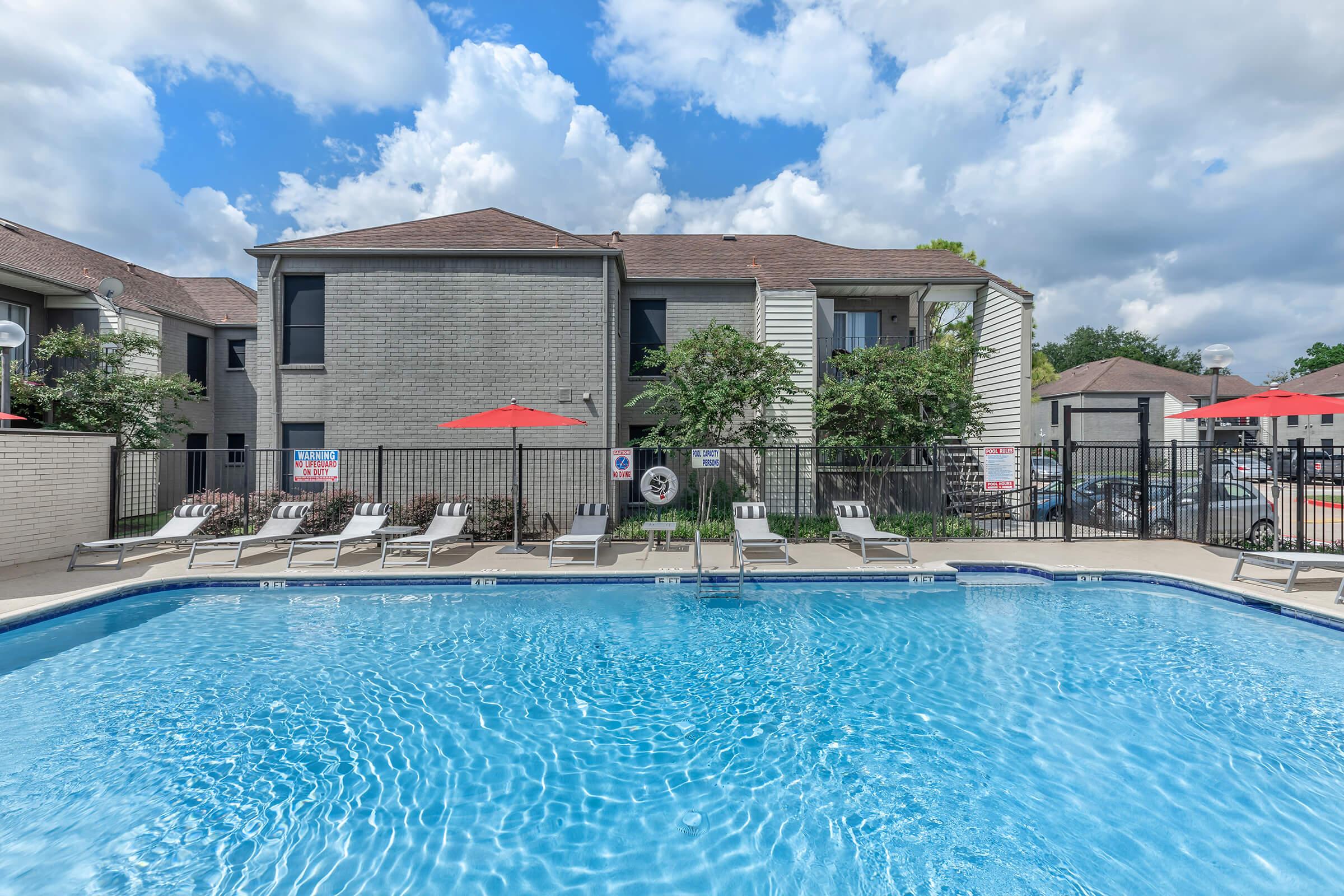 A clear blue swimming pool surrounded by lounge chairs and red umbrellas, with an apartment community in the background. The scene is set under a partly cloudy sky, adding a bright and inviting atmosphere to the outdoor space.