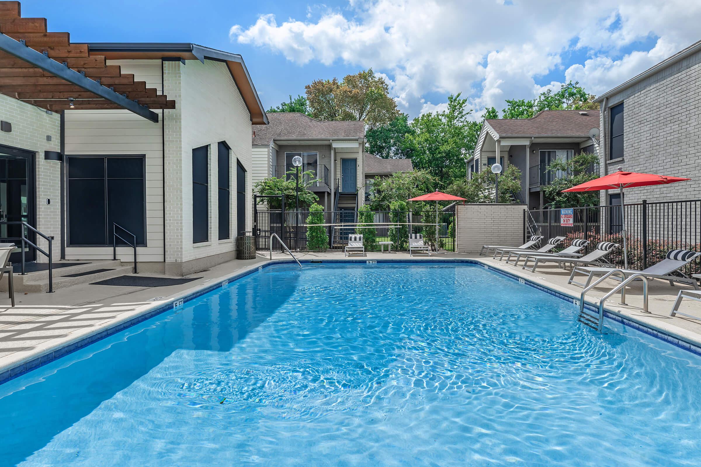 A serene swimming pool with clear blue water surrounded by lounge chairs. In the background, there are buildings with balconies and greenery. Red umbrellas provide shade, while a few trees and shrubs enhance the inviting atmosphere. The sky is partly cloudy, adding to the tranquil setting.