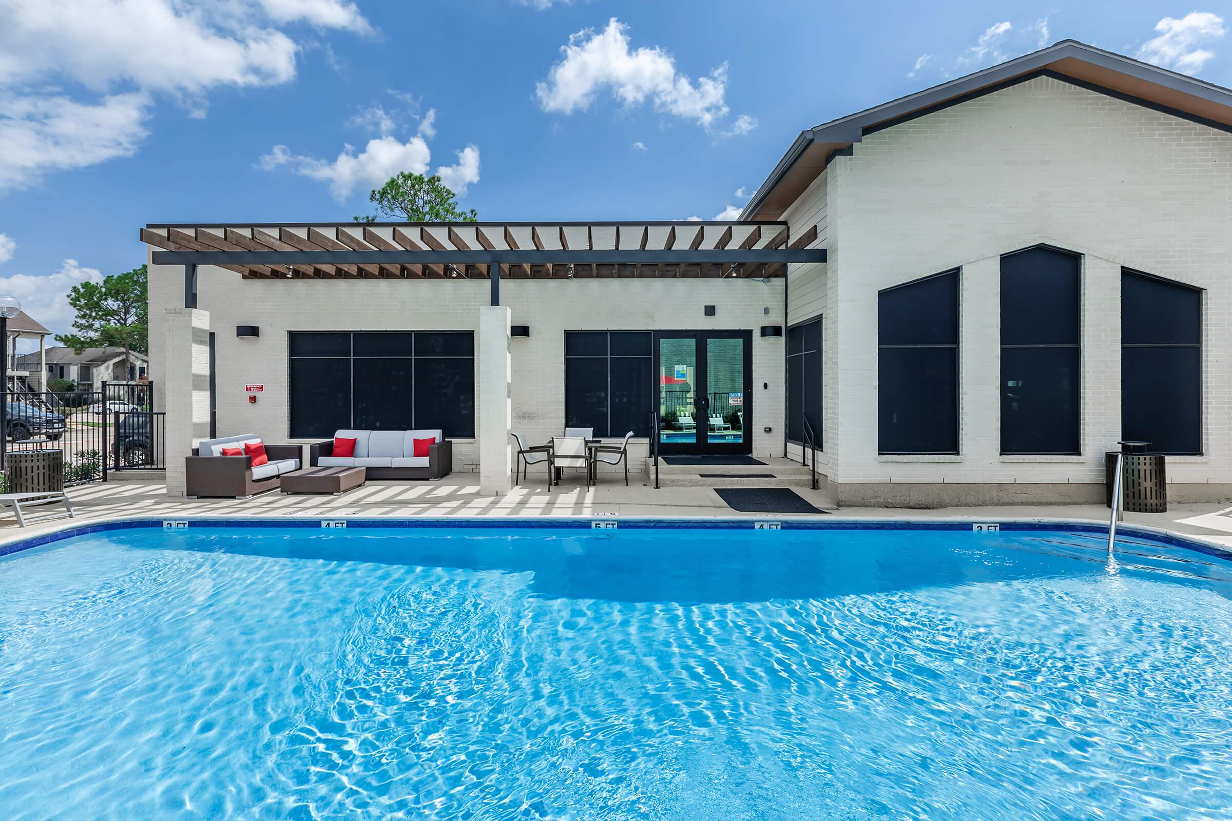 A bright, inviting swimming pool in front of a modern building. The pool features clear blue water, surrounded by lounge chairs and a small patio area with stylish furniture. The building has large windows and a shaded awning, set against a clear blue sky with a few fluffy clouds.