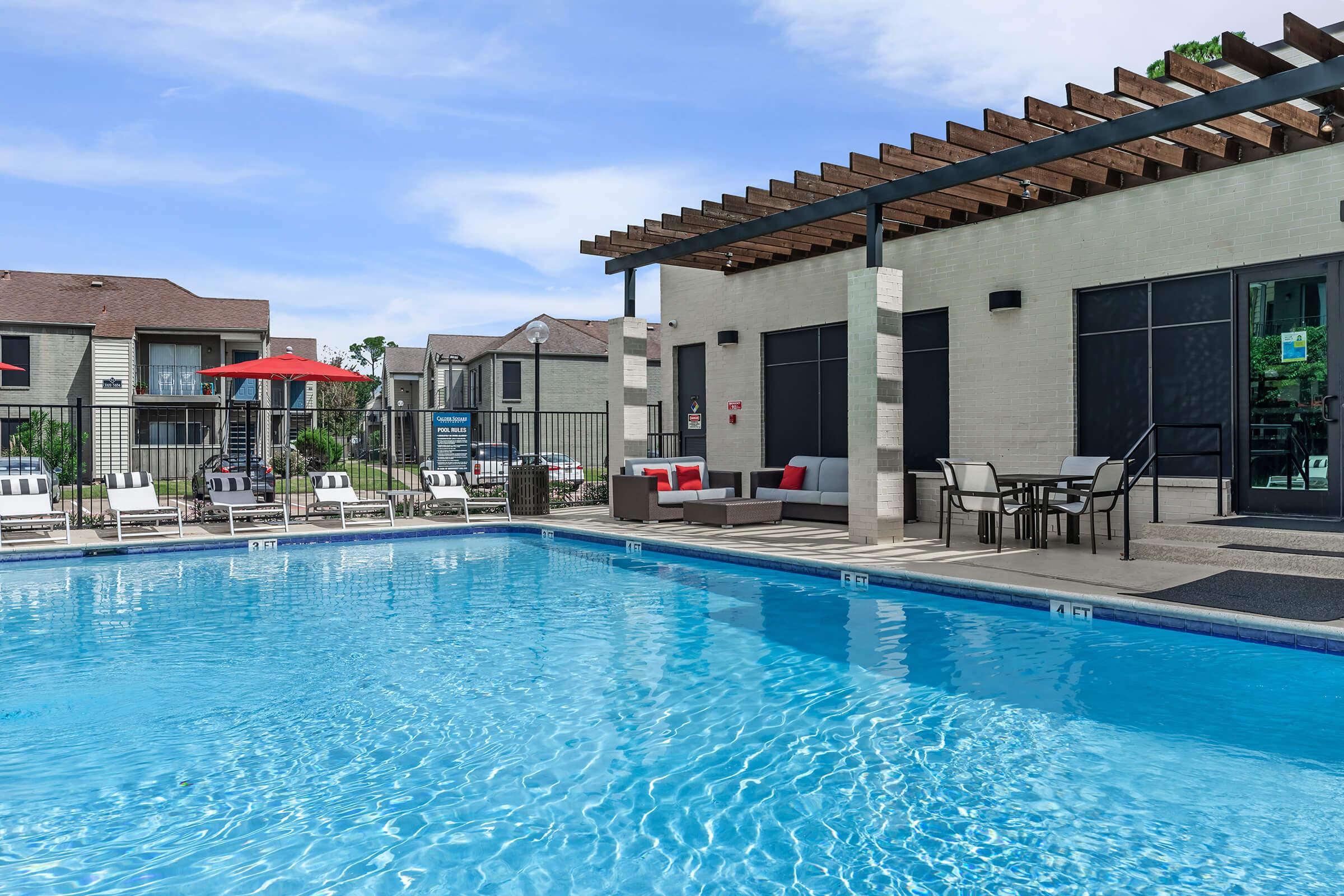 A clear swimming pool surrounded by lounge chairs and a shaded seating area. In the background, there are apartment buildings with balconies and red umbrellas. The scene is bright and inviting, featuring a blue sky and well-maintained landscape.