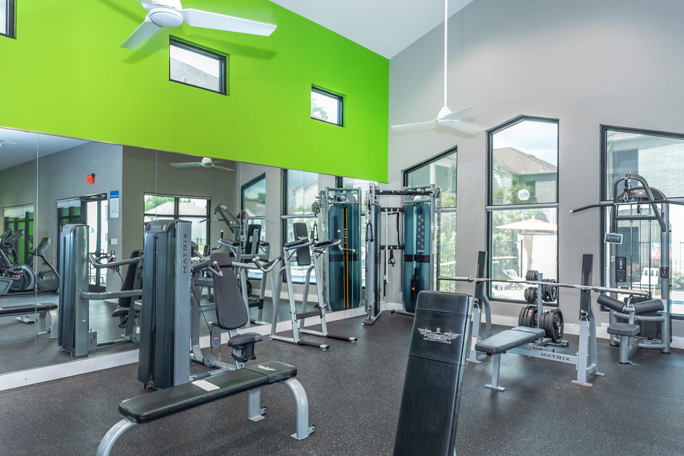 A modern gym interior featuring a workout area with various exercise machines, weights, and benches. Bright green accent wall and large windows allow natural light to illuminate the space. Fans are installed on the ceiling for ventilation. Mirrors line one wall, enhancing the open feel of the gym.