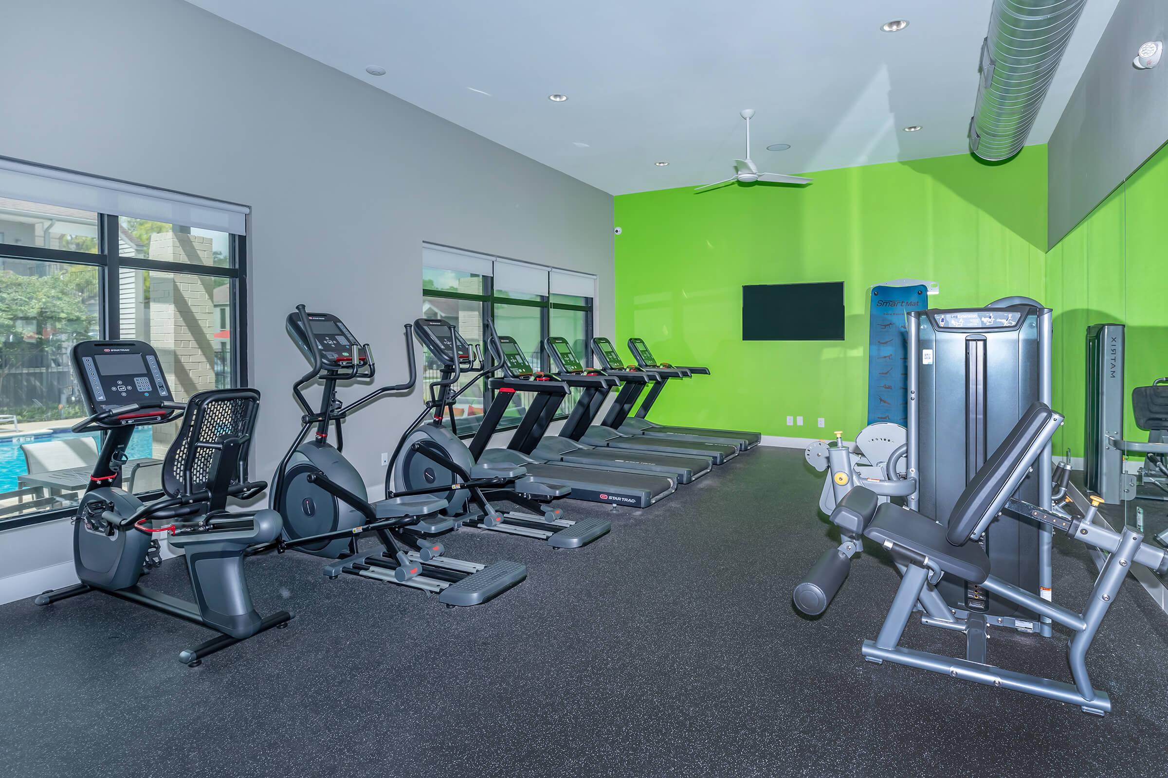 A modern gym interior featuring several cardio machines, including ellipticals and treadmills, along with a multi-use strength training machine. The space is well-lit with large windows and has a vibrant green accent wall, creating a motivating workout environment.