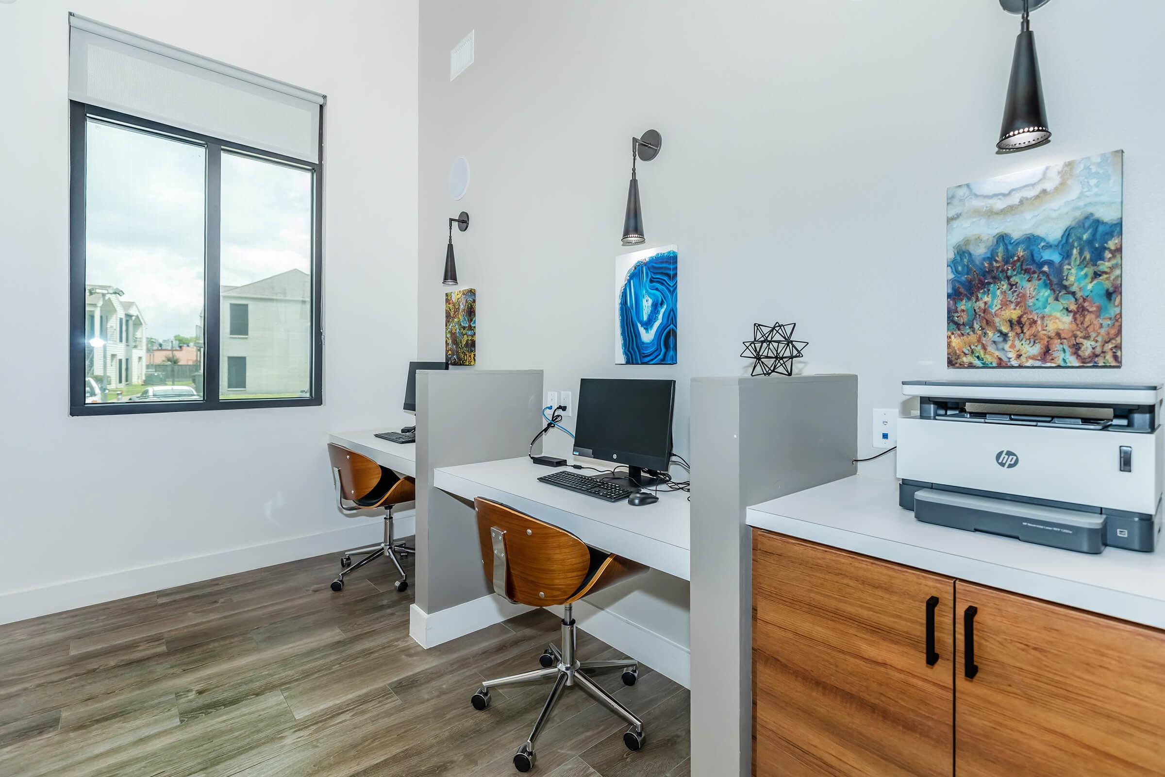 Two workstations with desktop computers are set up in a modern office space. Each workstation features a chair and decorative wall art nearby. A printer is placed on a cabinet against the wall, and a window provides natural light, revealing a view of buildings outside. The flooring is sleek and wooden.