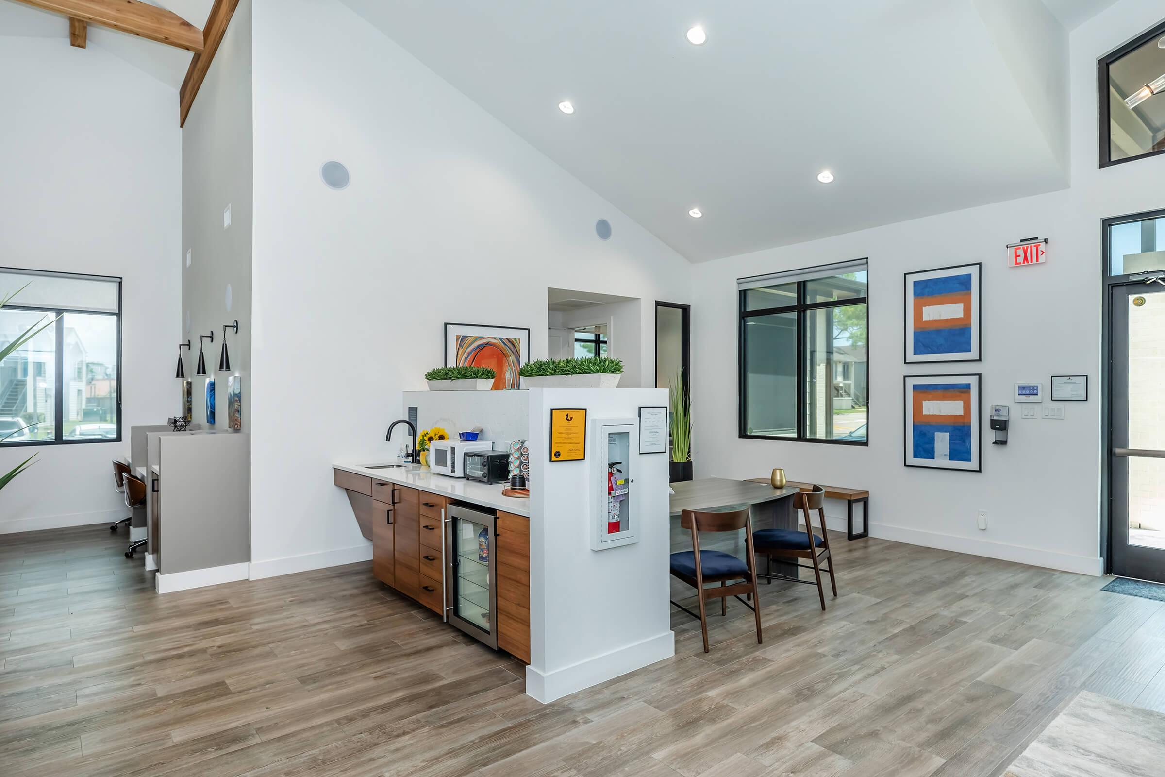 Modern interior of a communal space featuring a kitchenette with cabinets and appliances, a dining table with chairs, large windows letting in natural light, and abstract art on the walls. The space has wood accents and a high ceiling, creating an open and inviting atmosphere.