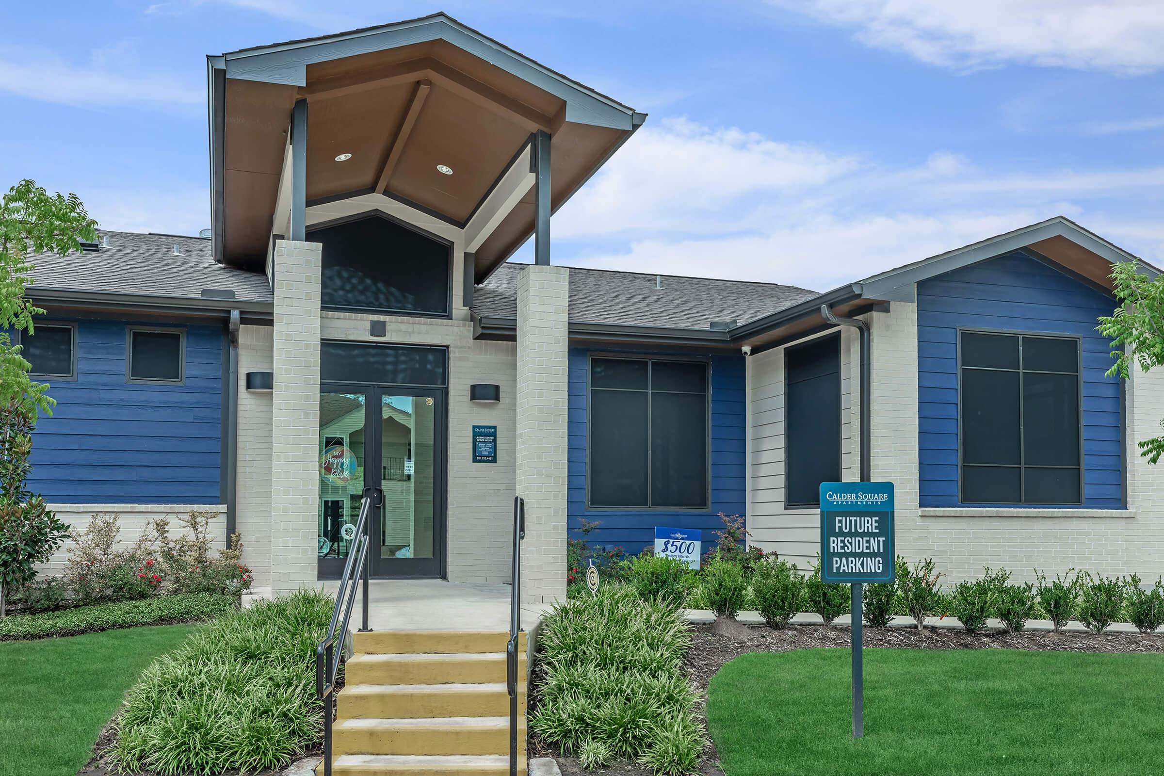 A modern apartment building entrance with a peaked roof and large windows. The facade features a mix of blue and white siding. Steps lead to the main door, and a sign indicates "Future Resident Parking." Lush green landscaping surrounds the building.