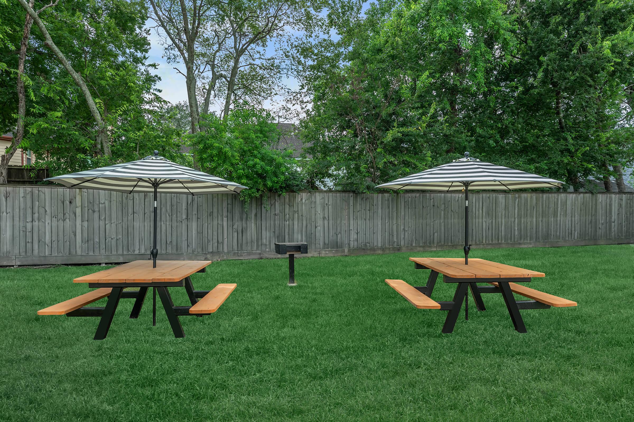 Two wooden picnic tables with black legs, each topped with a large striped umbrella, are set on a green lawn. Behind them, a wooden fence provides privacy, and a small grill is visible in the center. Lush trees line the background, creating a serene outdoor environment.