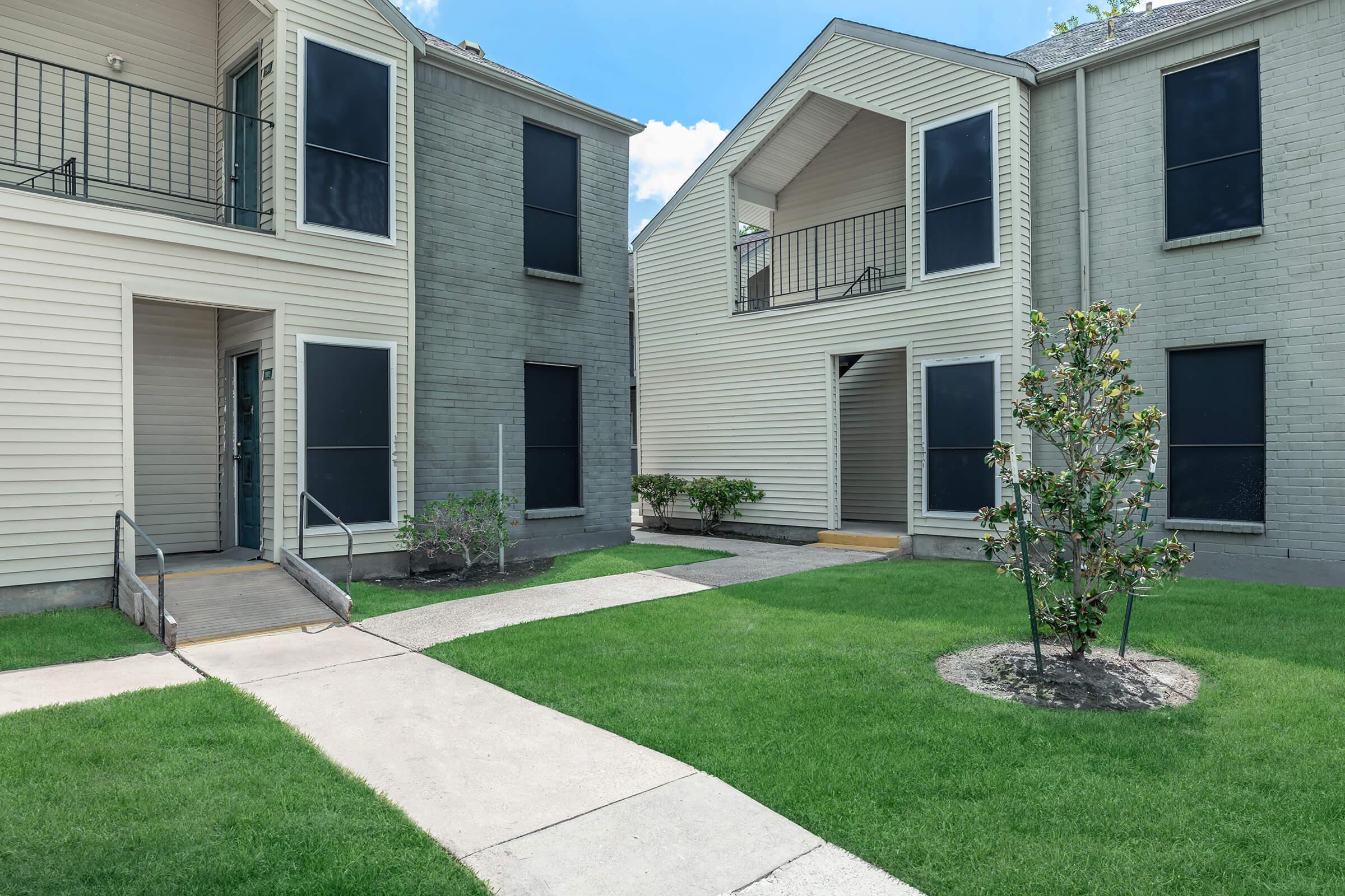 A view of two apartment buildings facing each other, featuring light-colored siding and black screen windows. A walkway lined with lush green grass connects the two buildings, and a small tree is planted in a landscaped area near the pavement. Bright blue sky and fluffy clouds are visible in the background.