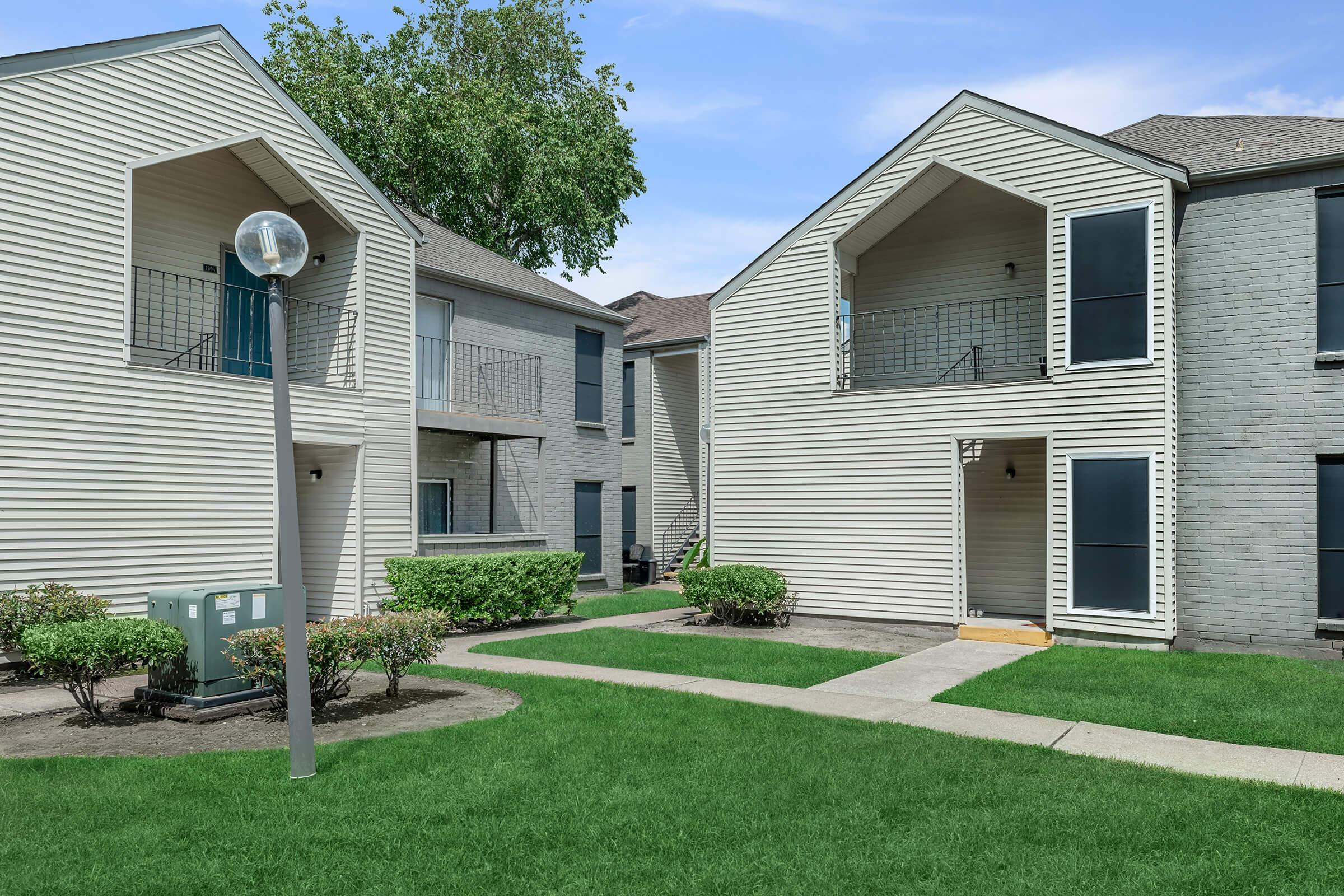 A view of a residential apartment community featuring two buildings with a landscaped courtyard. The buildings have light-colored siding, balconies, and large windows. A concrete walkway runs through the green lawn, leading to the entrances of the apartments. Decorative bushes and a light post are visible in the foreground.