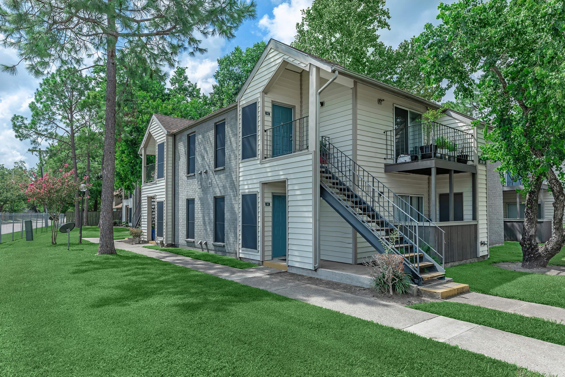 A two-story residential building with a light-colored exterior, featuring balconies and a metal staircase. Surrounding the building is a green lawn and trees. A sidewalk runs alongside the building, with a street visible in the background. The sky is partly cloudy, creating a bright atmosphere.