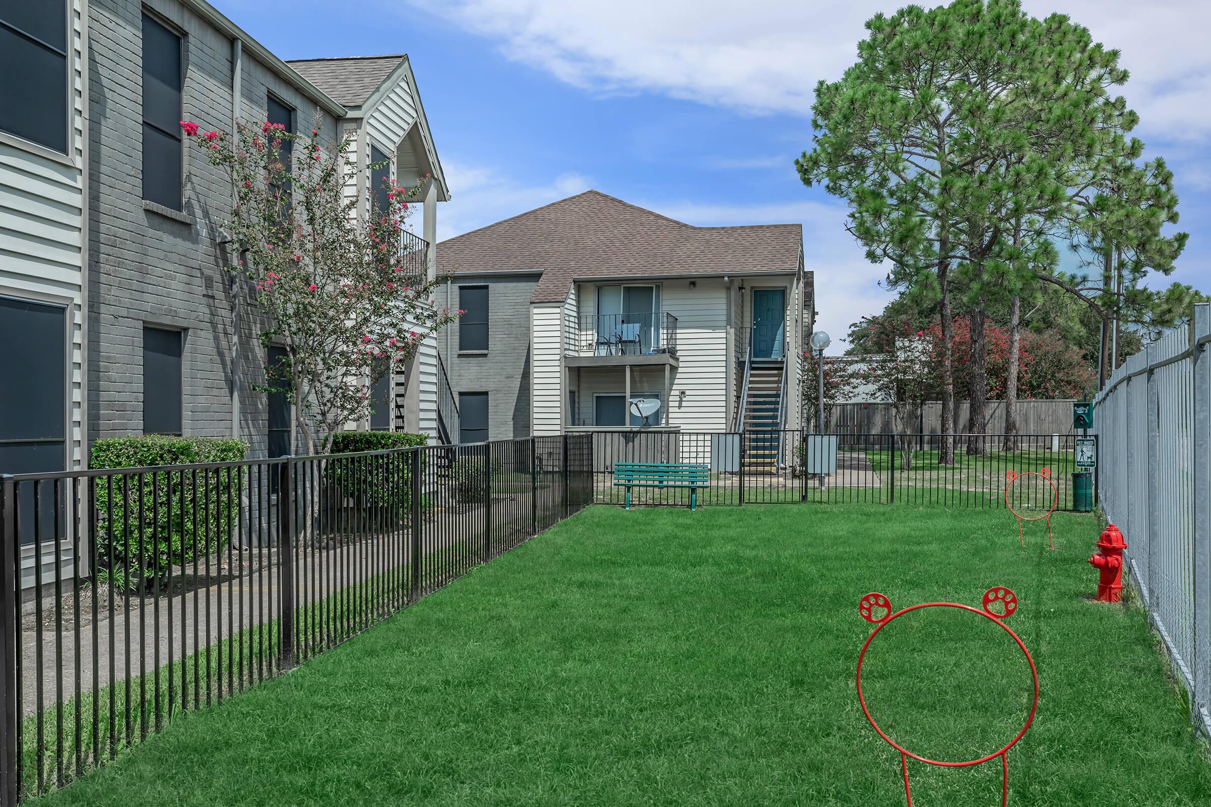 A well-maintained apartment community surrounded by green grass, featuring two buildings with balconies. A fenced area includes a red fire hydrant and a playful dog agility course with a hoop. Trees and shrubs provide greenery, and the sky is partly cloudy.