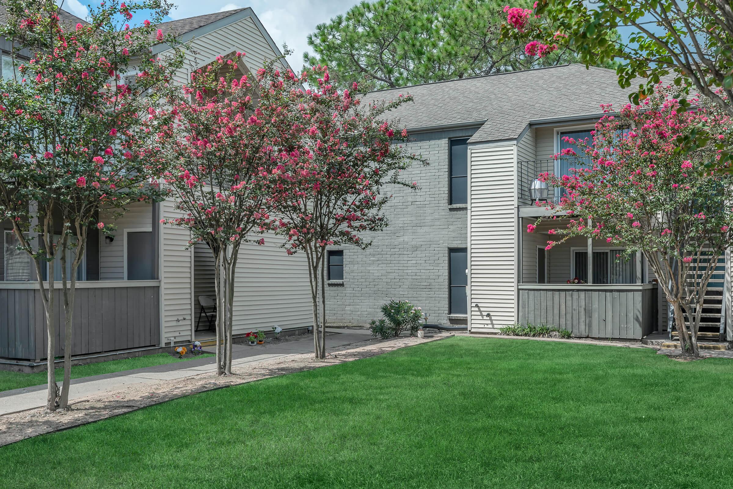 A well-maintained residential complex featuring two-story buildings with white and beige siding. Vibrant pink flowering trees are situated in the foreground, enhancing the lush green lawn. The scene is bright and inviting, with a clear blue sky in the background.