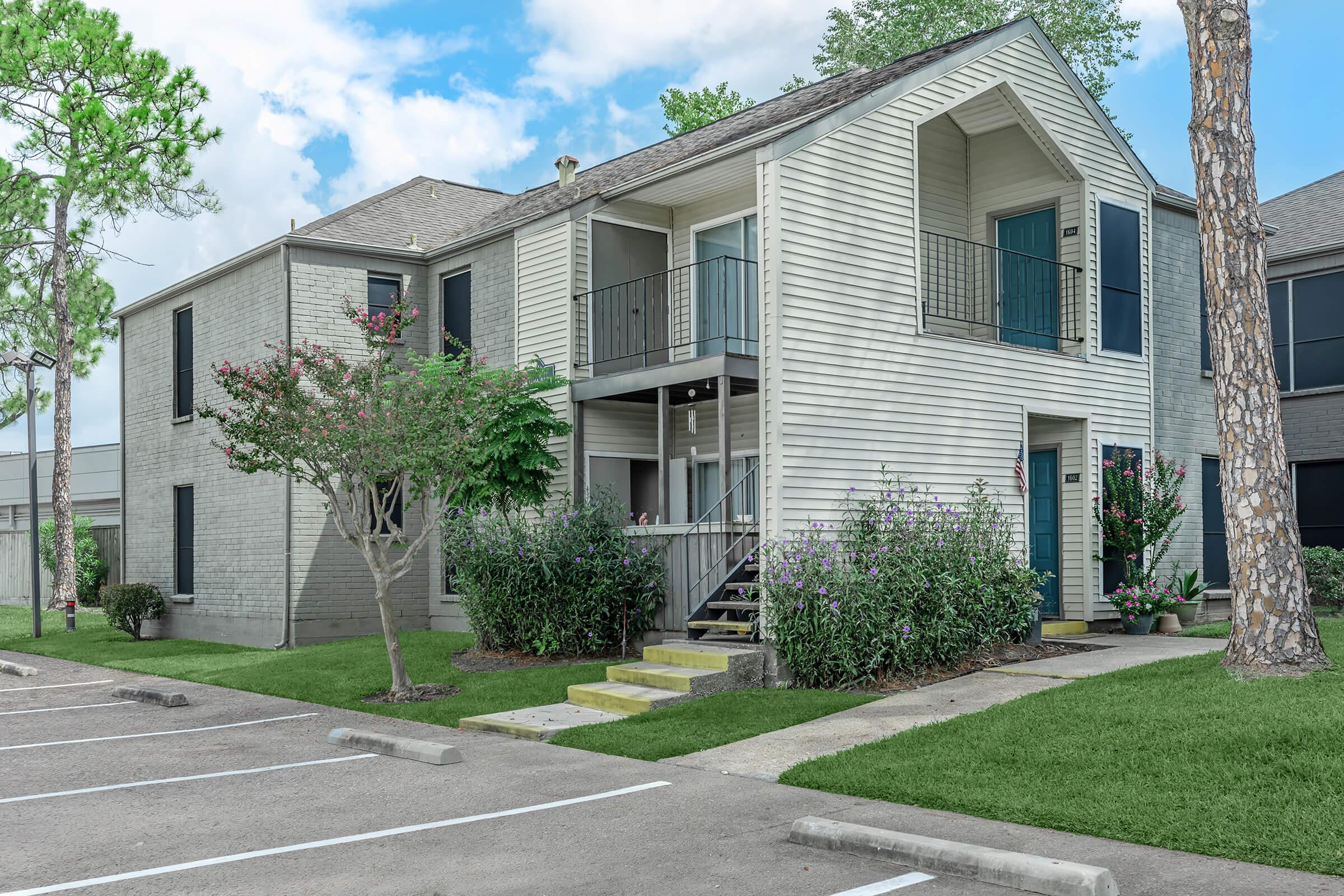 A two-story residential building with a light-colored wooden façade, featuring balconies on the upper level. Lush greenery and flowering shrubs surround the stairs leading to the entrance. The parking lot is visible in the foreground, with several empty parking spaces. Blue skies with scattered clouds are in the background.