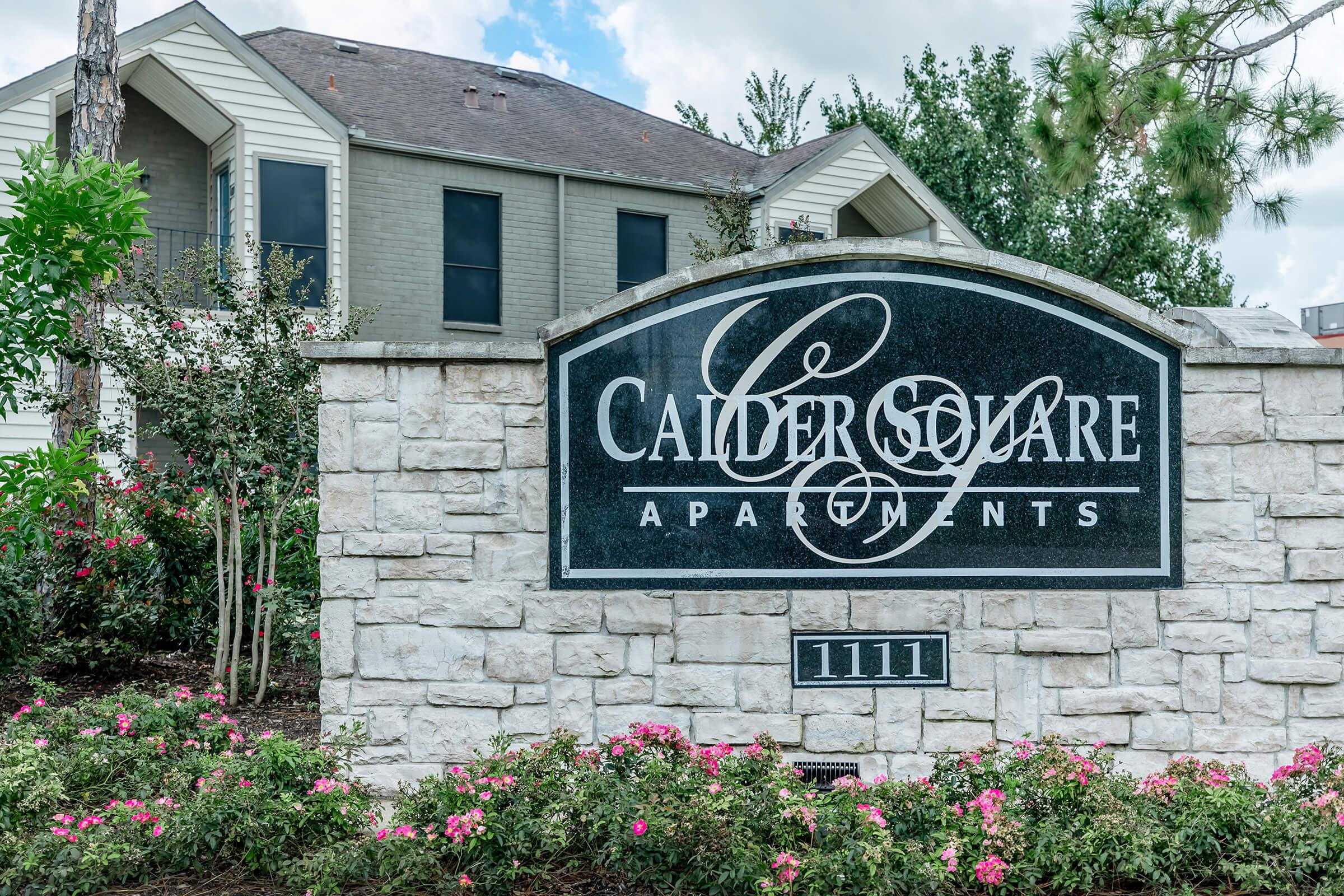 Sign for Calder Square Apartments, featuring the name "Calder Square" in elegant lettering. The sign is made of dark stone, framed by light-colored stone, with decorative flowers and greenery surrounding it. In the background, a residential building can be seen.