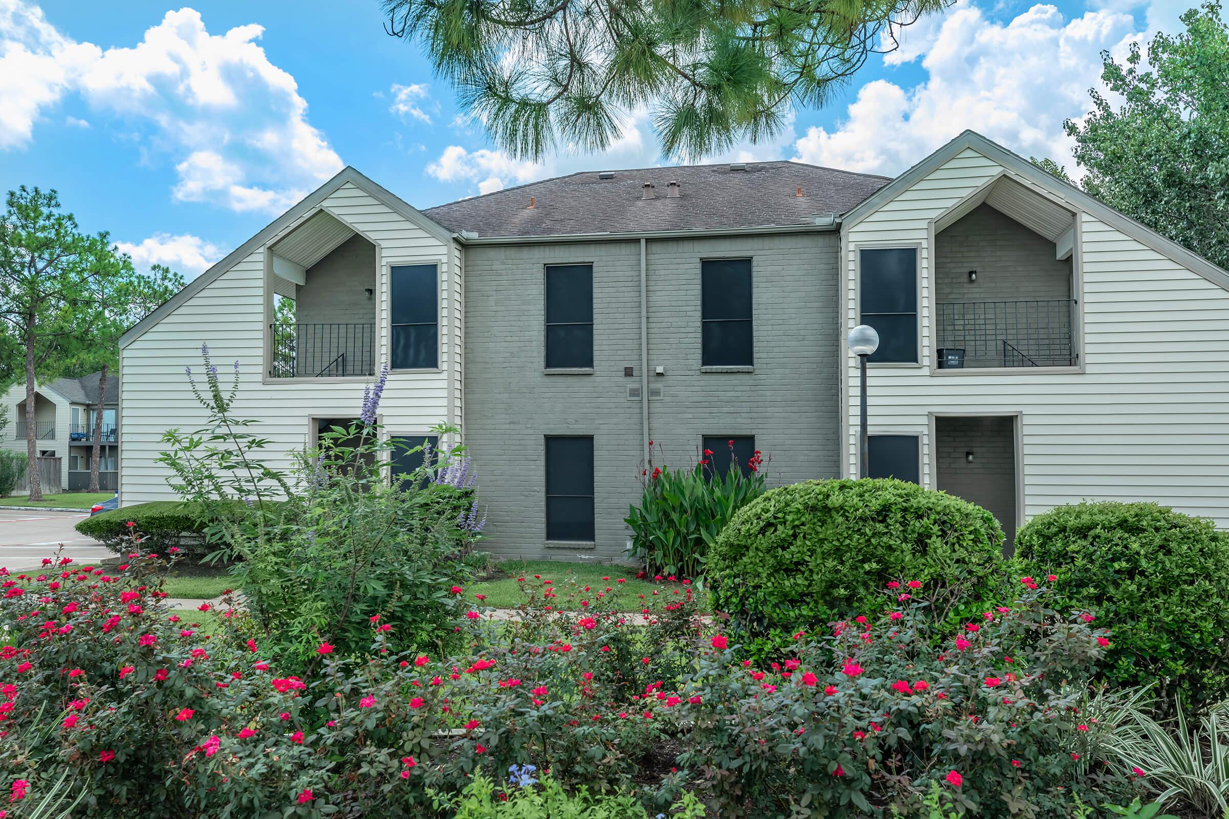 Two-story apartment building with a light gray exterior and triangular rooflines. Surrounding the building, there are lush green bushes and vibrant flower beds featuring red and pink blooms. The sky above is blue with fluffy white clouds, contributing to a serene residential atmosphere.