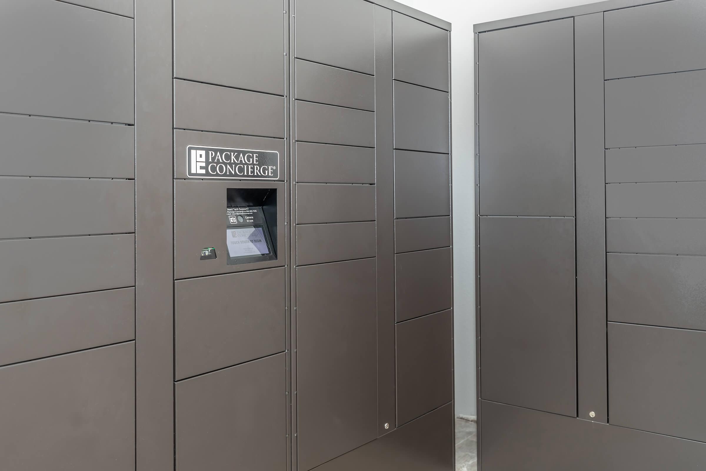 A row of gray package lockers with a central touch screen display labeled "PACKAGE CONCIERGE." The lockers are designed for secure package delivery and retrieval, providing a modern solution for receiving packages in residential or commercial buildings.