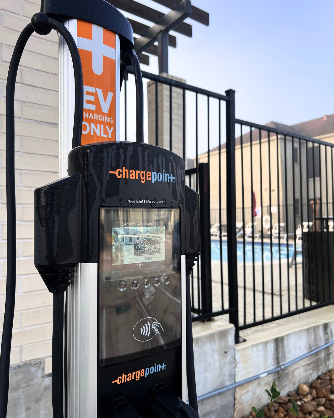 Electric vehicle (EV) charging station labeled "ChargePoint" with a display screen. The charger is located near a fenced area with a swimming pool visible in the background. The sign indicates that it is for EV charging only.