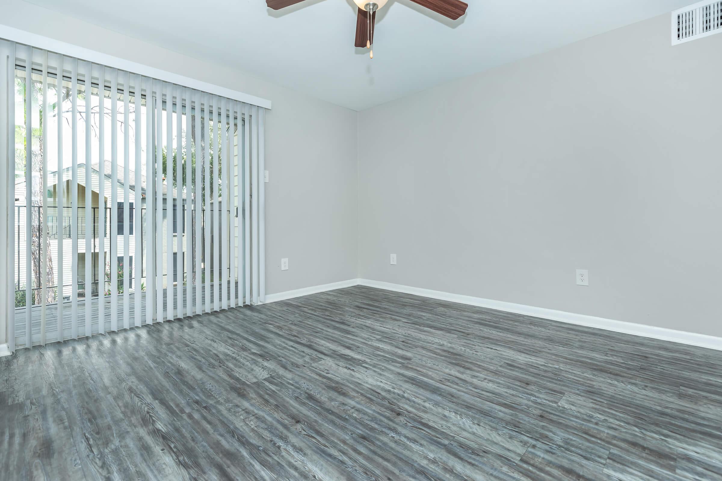 A spacious, empty room with light gray walls and a ceiling fan. There are sliding glass doors with vertical blinds leading to a balcony, allowing natural light to enter. The floor features dark wood laminate, enhancing the modern look of the space.