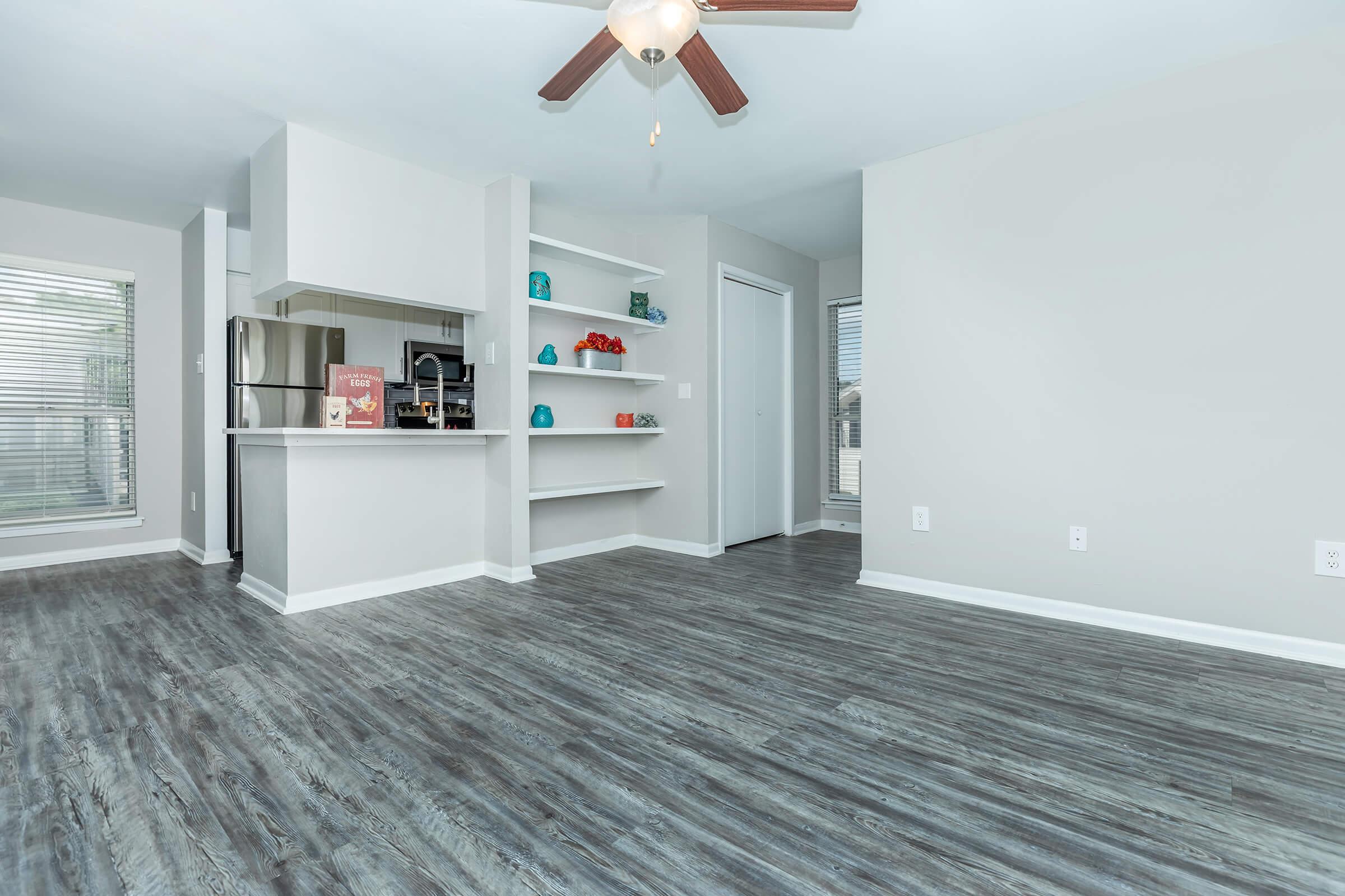 Bright and modern living space featuring gray wood-style flooring, a ceiling fan, and an open kitchen area with stainless steel appliances. The walls are painted in a soft light color, and shelves display colorful decorative items. Large windows let in natural light, creating an inviting atmosphere.