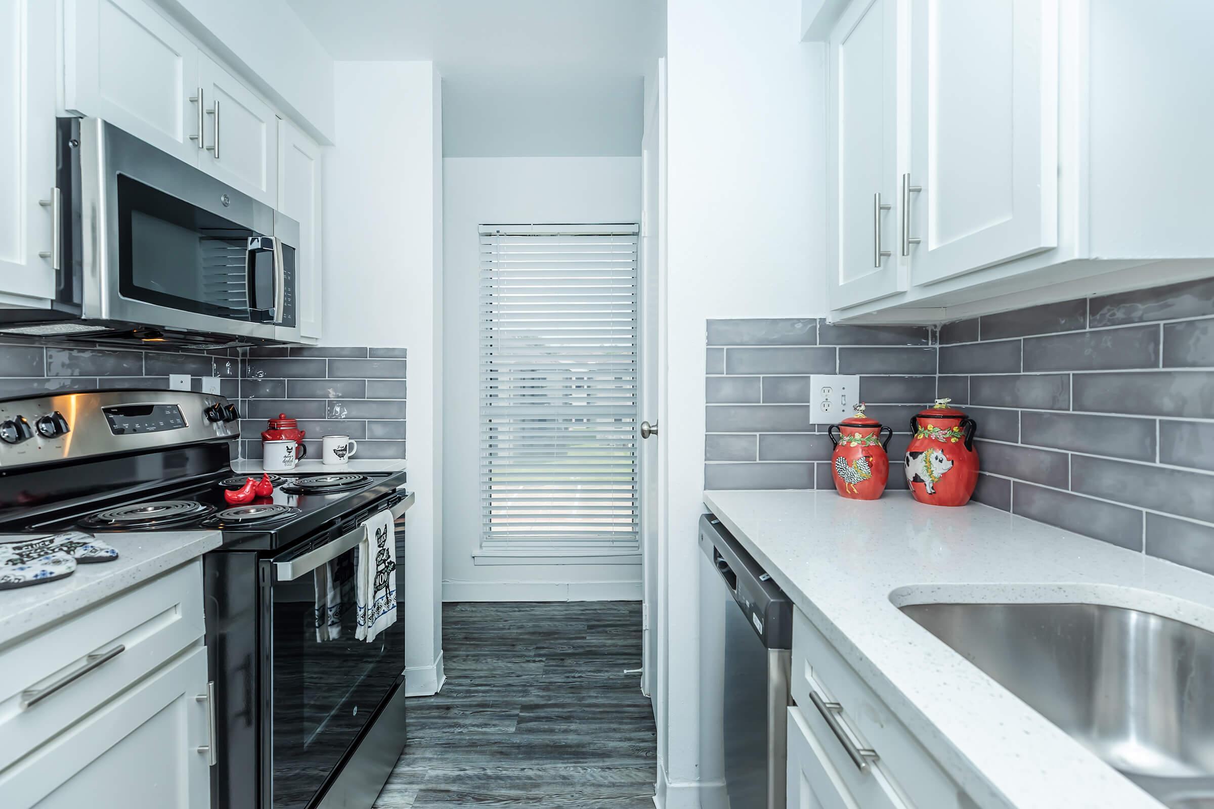 Modern kitchen with white cabinets and gray tile backsplash. Features stainless steel appliances, including a microwave and oven. A view of a window with blinds provides natural light, and decorative red jars are placed on the countertop. The flooring is dark and contrasts with the light-colored cabinets.