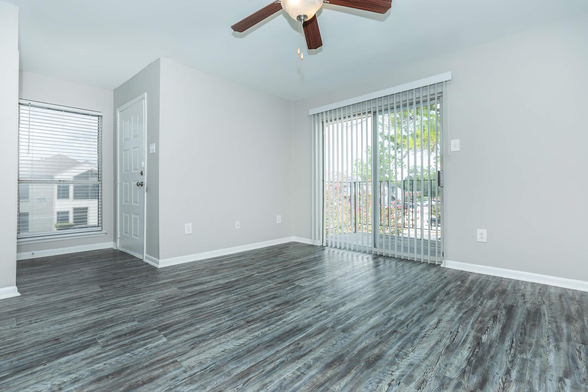 A bright, empty room with gray walls and dark laminate flooring. It features a ceiling fan and large sliding glass doors with vertical blinds leading to a balcony. A window with white blinds is visible on the left, allowing natural light to enter the space. The room is ready for furniture placement.