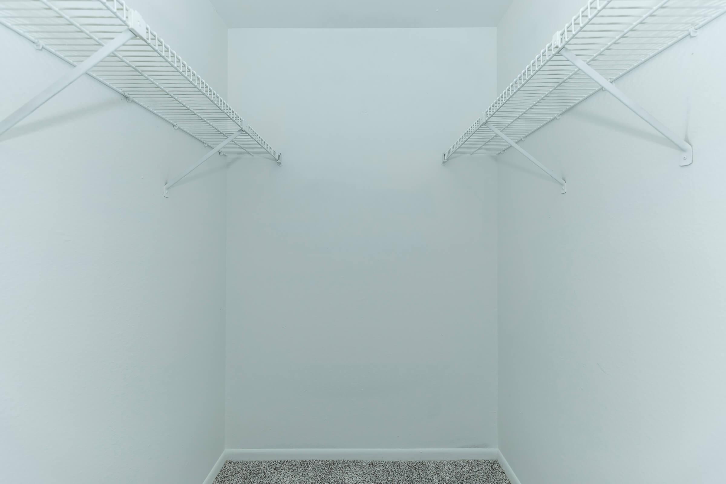 A clean, empty closet with white walls and two wire shelving units on opposite sides. The floor is covered with a light-colored textured material, and the space is well-lit, emphasizing its simplicity and emptiness.