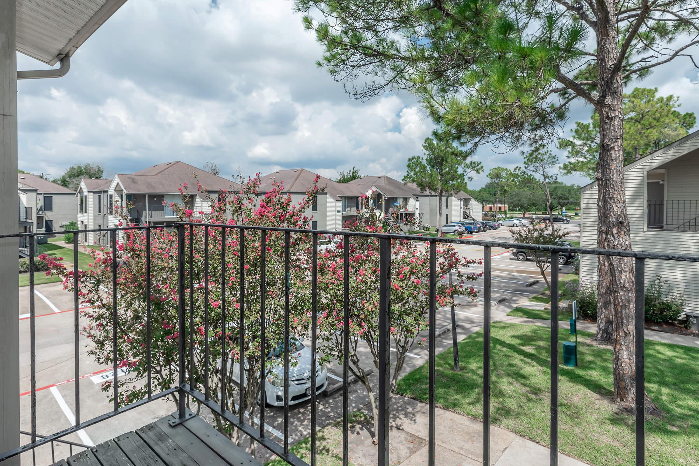 View from a balcony overlooking a residential area. The scene includes flowering trees, parked cars, and several apartment buildings in the background under a partly cloudy sky. Lush green grass and landscaping are visible in the foreground, creating a peaceful suburban atmosphere.