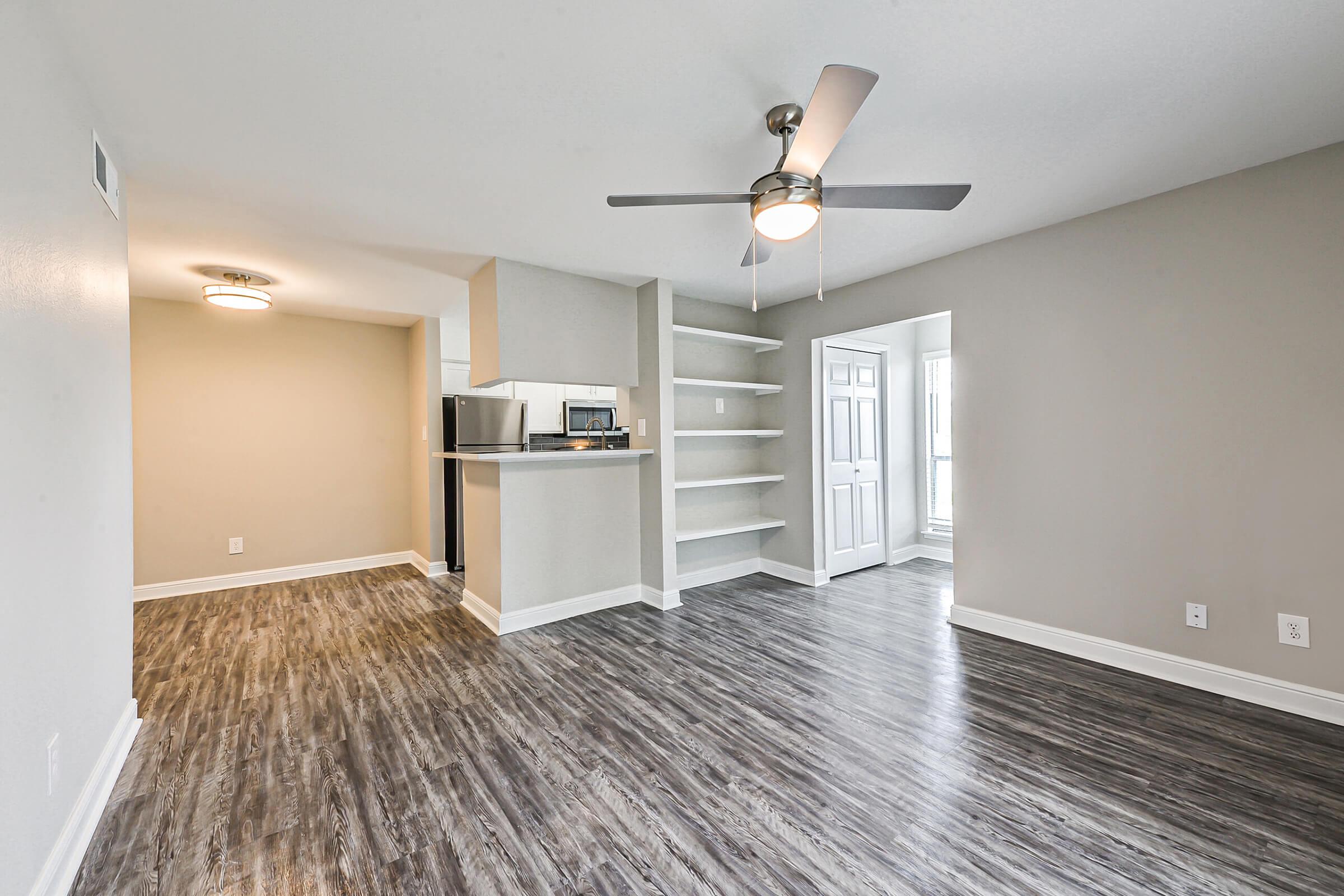 A spacious living area featuring light gray walls, a ceiling fan, and modern laminate flooring. On one side, there's a small kitchen area with a bar counter, and on the other side, a doorway leading to another room. The overall ambiance is bright and inviting, ideal for a cozy home setting.