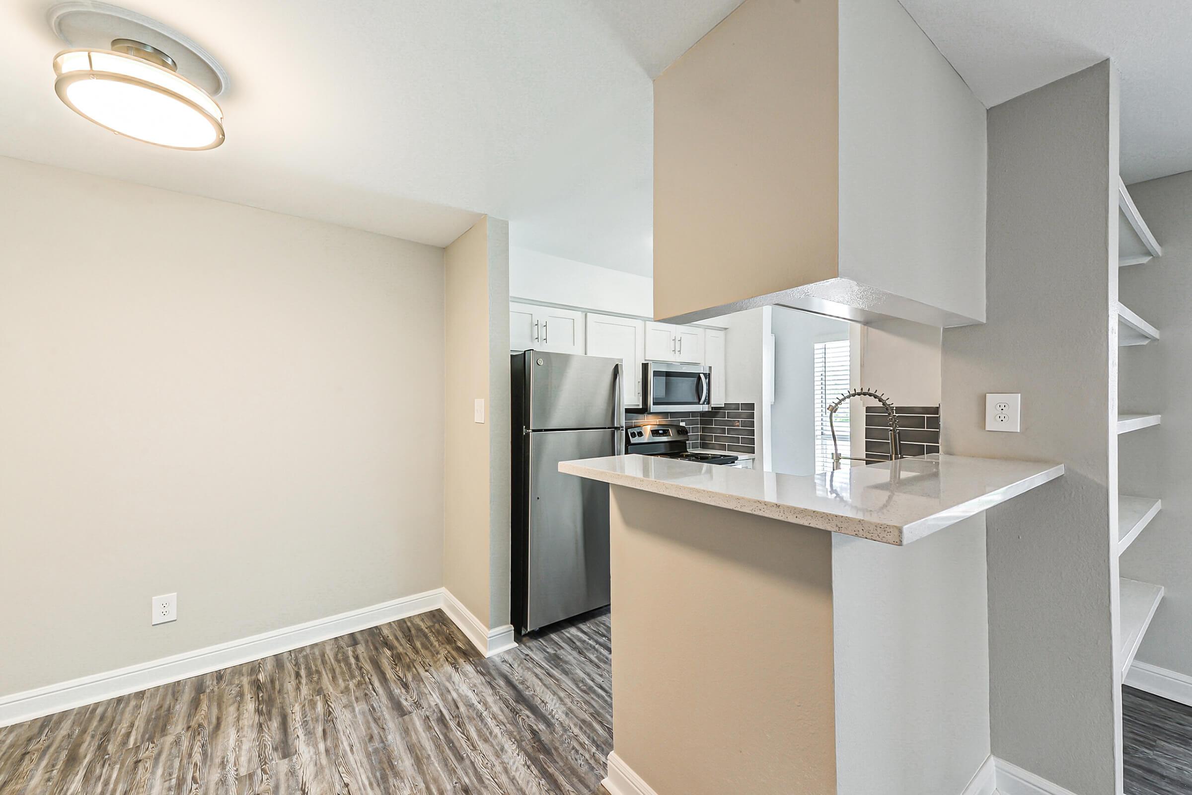 A modern kitchen with a light fixture overhead, featuring stainless steel appliances including a refrigerator and microwave. The space includes a bar-height countertop with a sink, surrounded by light grey walls and wooden flooring, creating a clean and contemporary look.