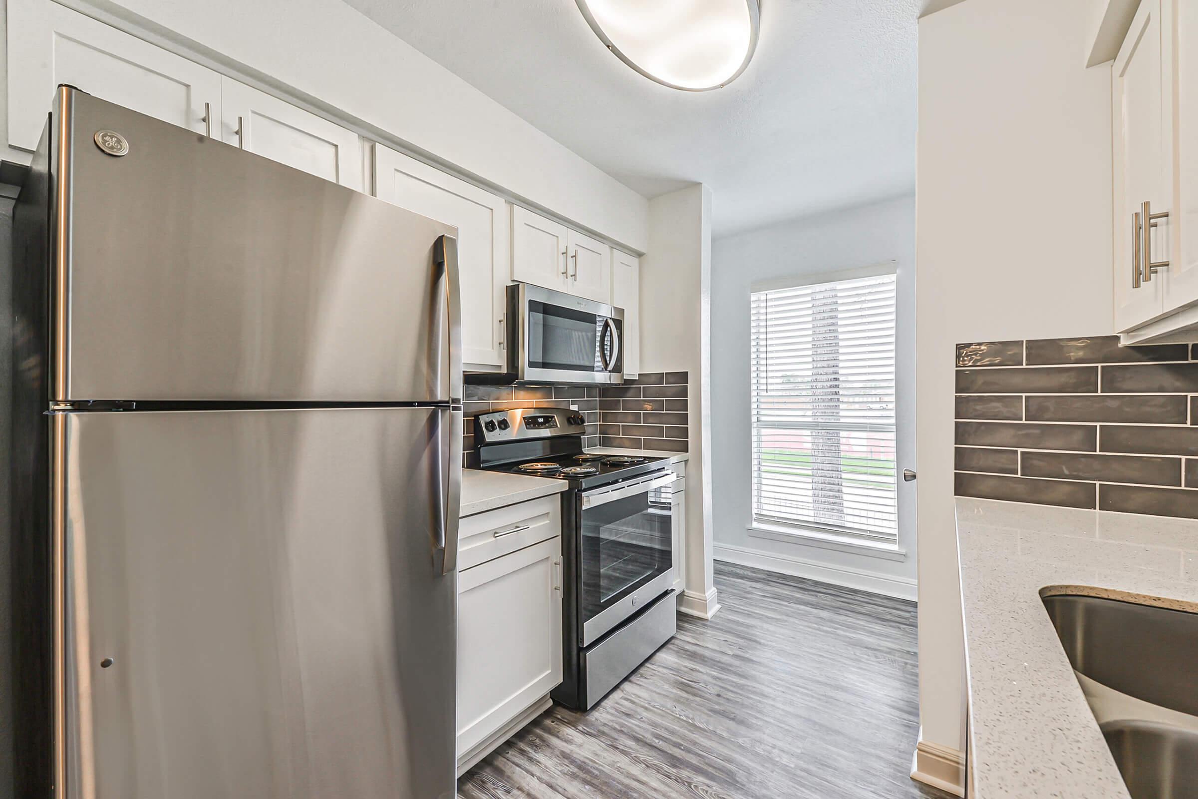 A modern kitchen featuring stainless steel appliances including a refrigerator, oven, and microwave. The cabinets are white, and the backsplash is composed of dark tiles, creating a sleek contrast. Natural light filters through a window with blinds, highlighting the wood-look flooring.