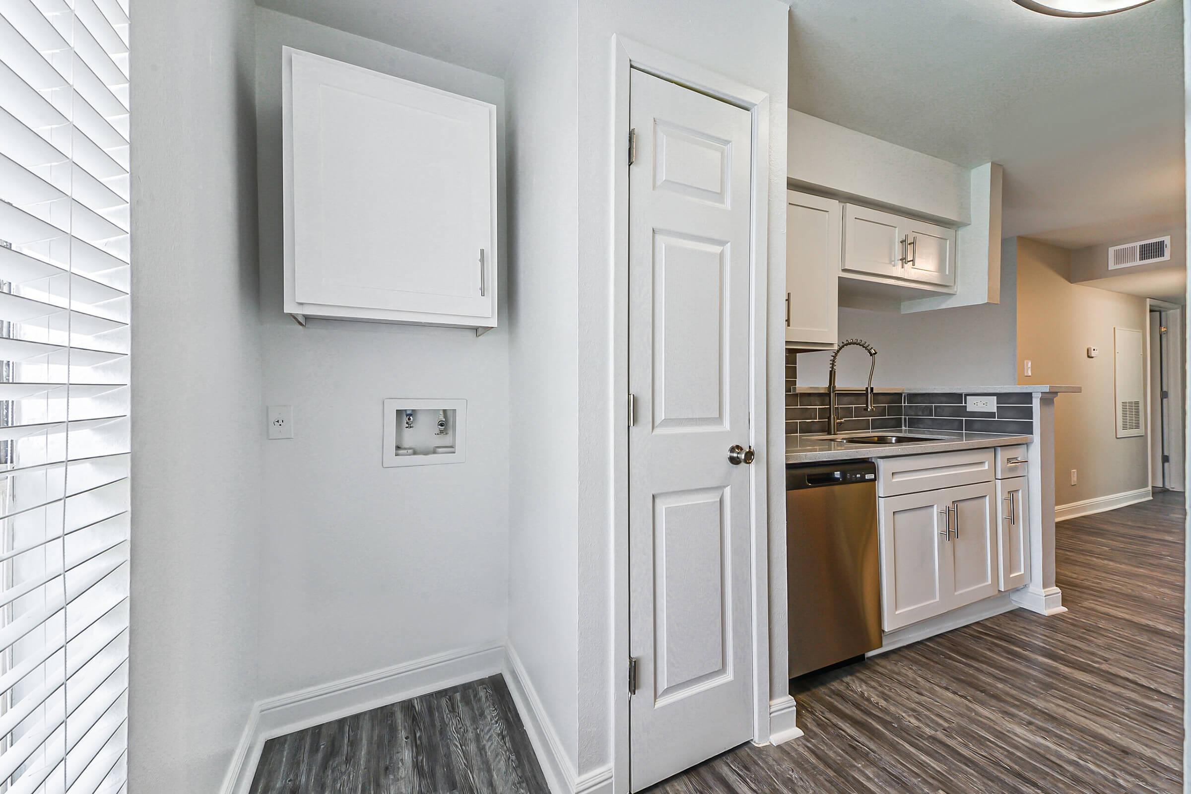 A modern kitchen featuring white cabinets, silver appliances, and a stylish backsplash. There is a dishwasher, a closed door leading to another room, and a laundry nook with electrical connections. Natural light streams in through a nearby window with blinds, enhancing the contemporary look of the space.