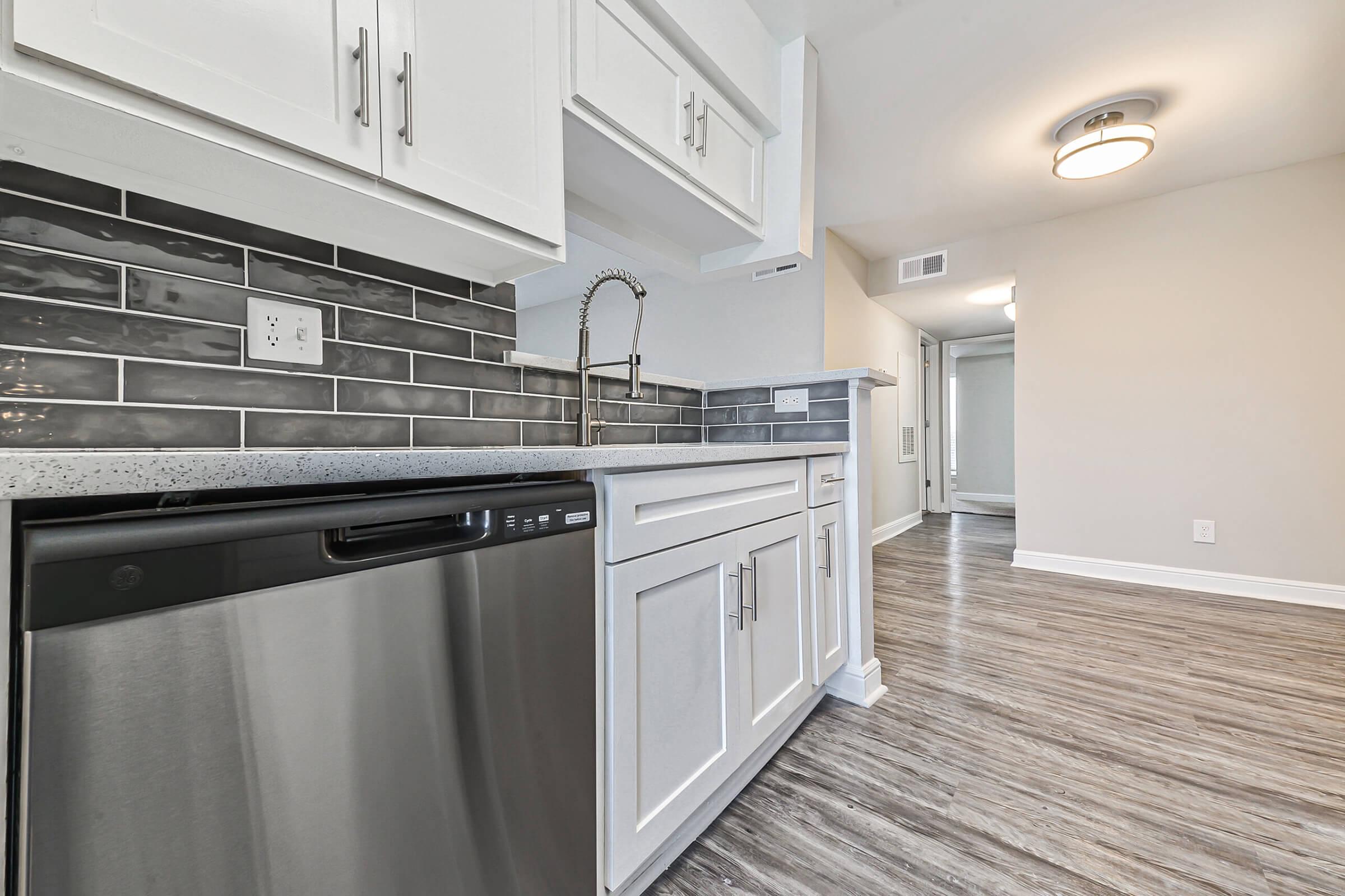 Modern kitchen featuring white cabinetry, a stainless steel dishwasher, and a gray tiled backsplash. The counter is a light granite with an integrated sink and a sleek faucet. The flooring is a light brown wood-like laminate. A bright ceiling light fixture illuminates the space, with a doorway leading to another room.