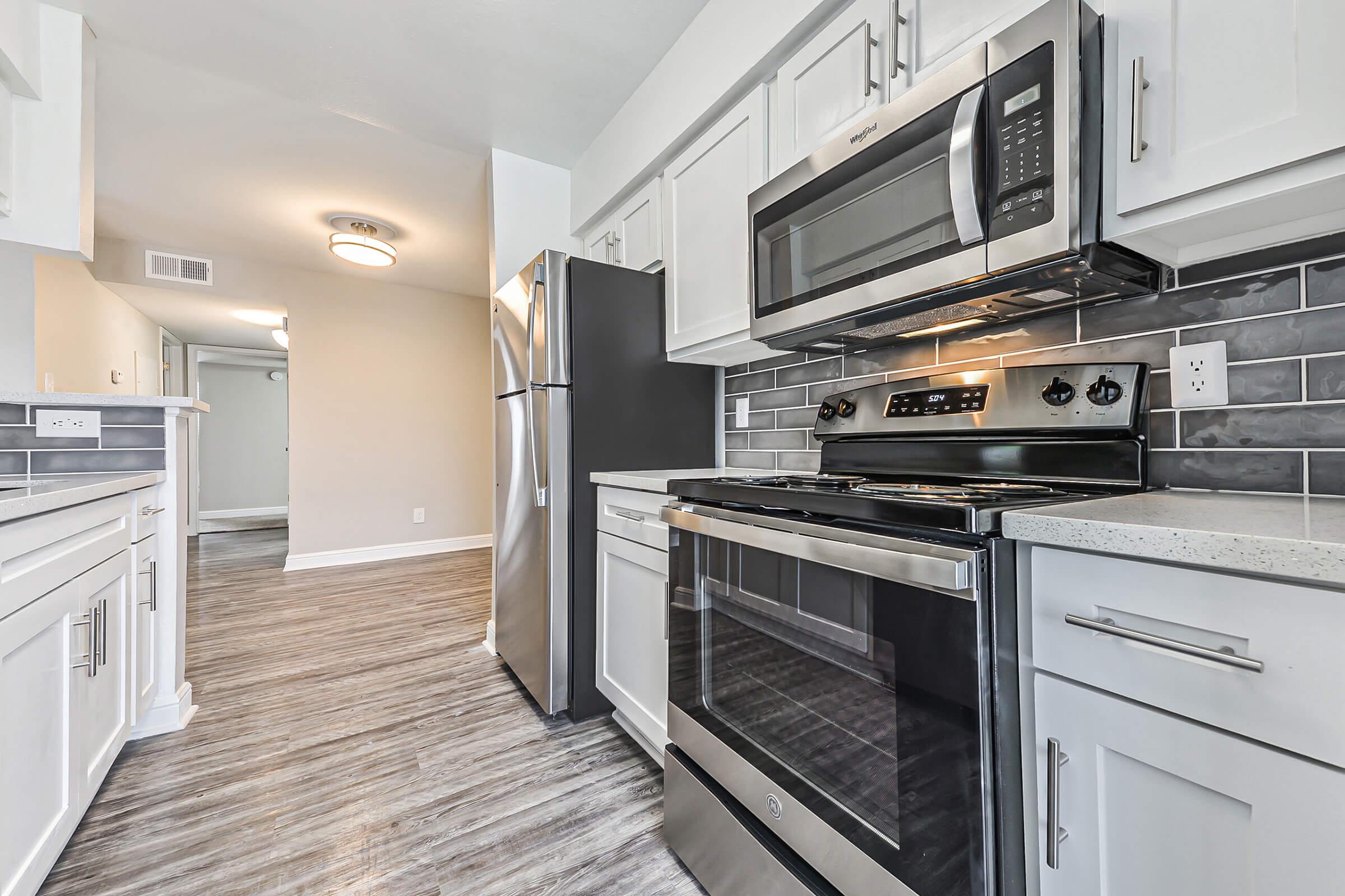 Modern kitchen featuring stainless steel appliances, including a stove, microwave, and refrigerator. The cabinetry is white, and the countertops are light-colored with a gray tile backsplash. The flooring is in a wood-like finish, adding warmth to the space. Soft lighting enhances the bright, clean atmosphere.