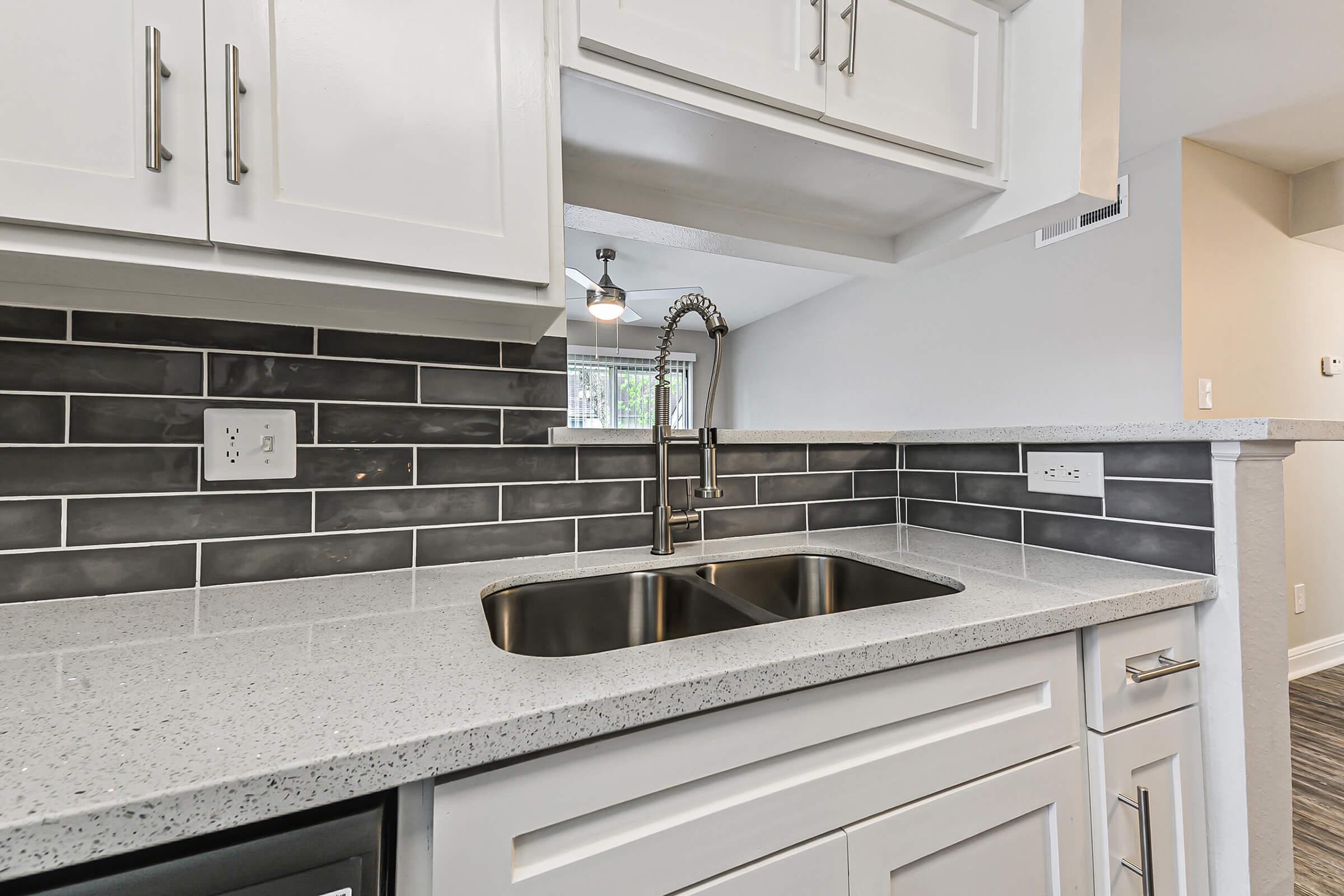 Modern kitchen with a sleek design featuring white cabinetry, a gray and black tiled backsplash, and a double sink. The countertop has a light speckled finish, and there is a stylish pull-down faucet. Natural light filters in through a nearby window, enhancing the contemporary feel of the space.