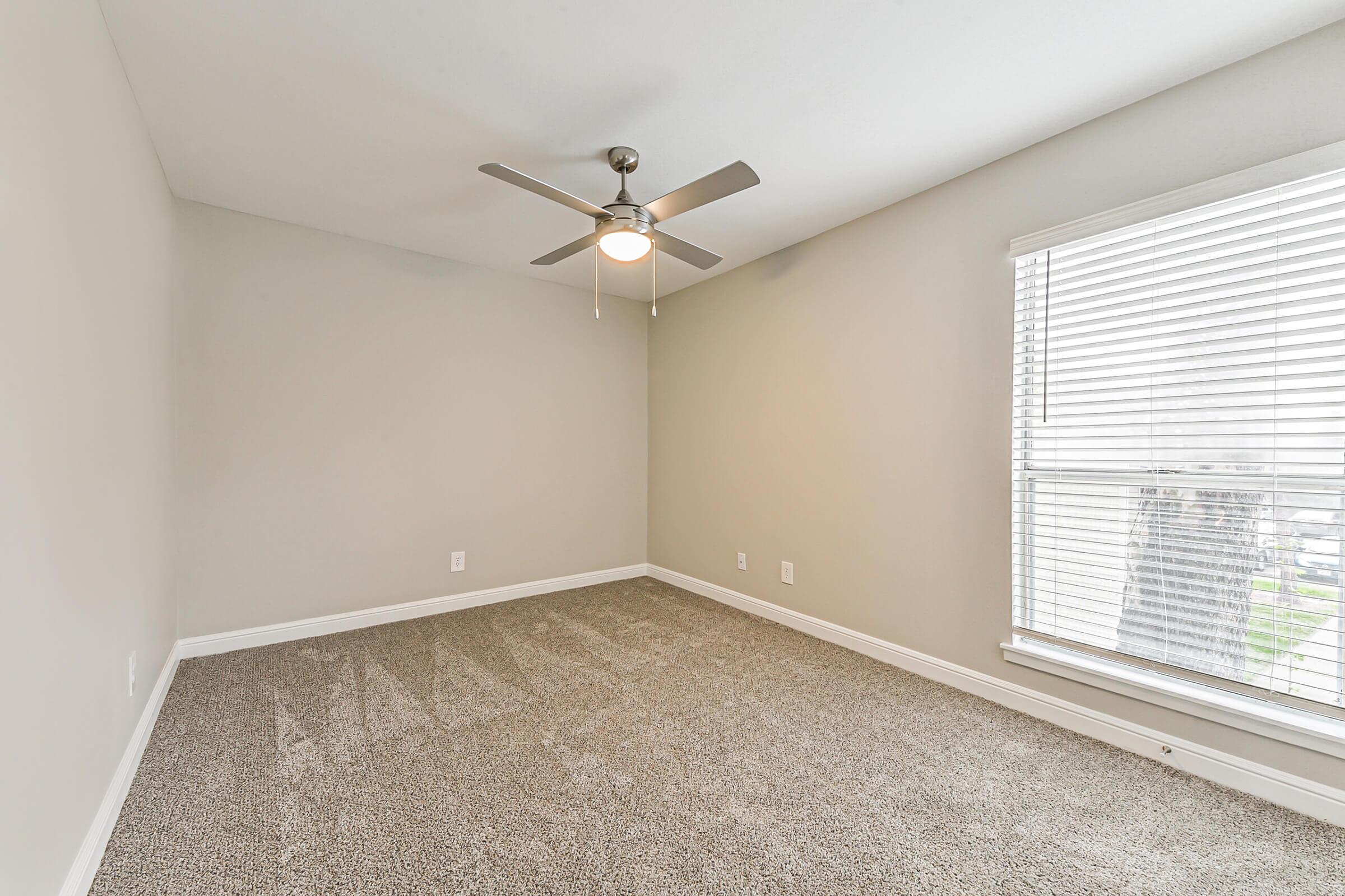 A minimalistic, empty room featuring light gray walls and carpet. It includes a ceiling fan with a light fixture and a window with closed blinds allowing natural light to enter, illuminating the space. The room is void of furniture, creating a spacious and clean atmosphere.