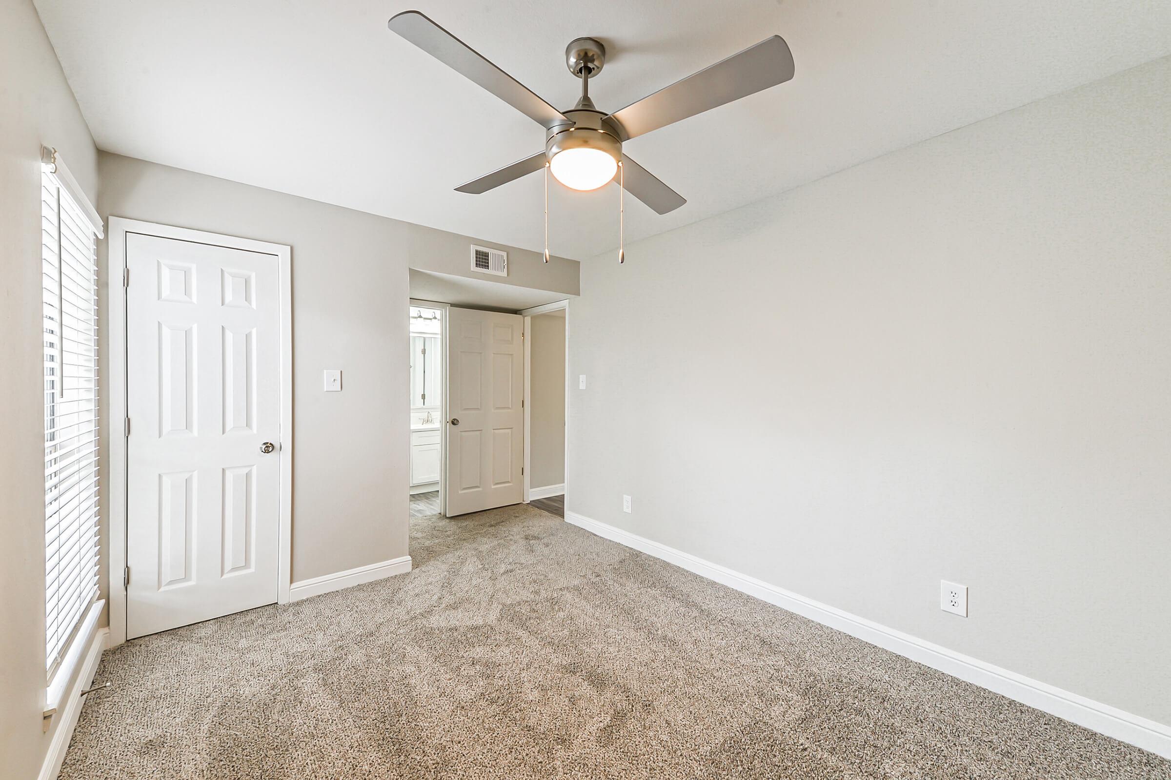 A well-lit, neutral-colored bedroom featuring plush, light gray carpet. It has a ceiling fan with a light fixture and two closed doors leading to other rooms. Windows on the left provide natural light, while the walls are painted in a soft shade. The room has a clean, modern aesthetic, ideal for personal or guest use.