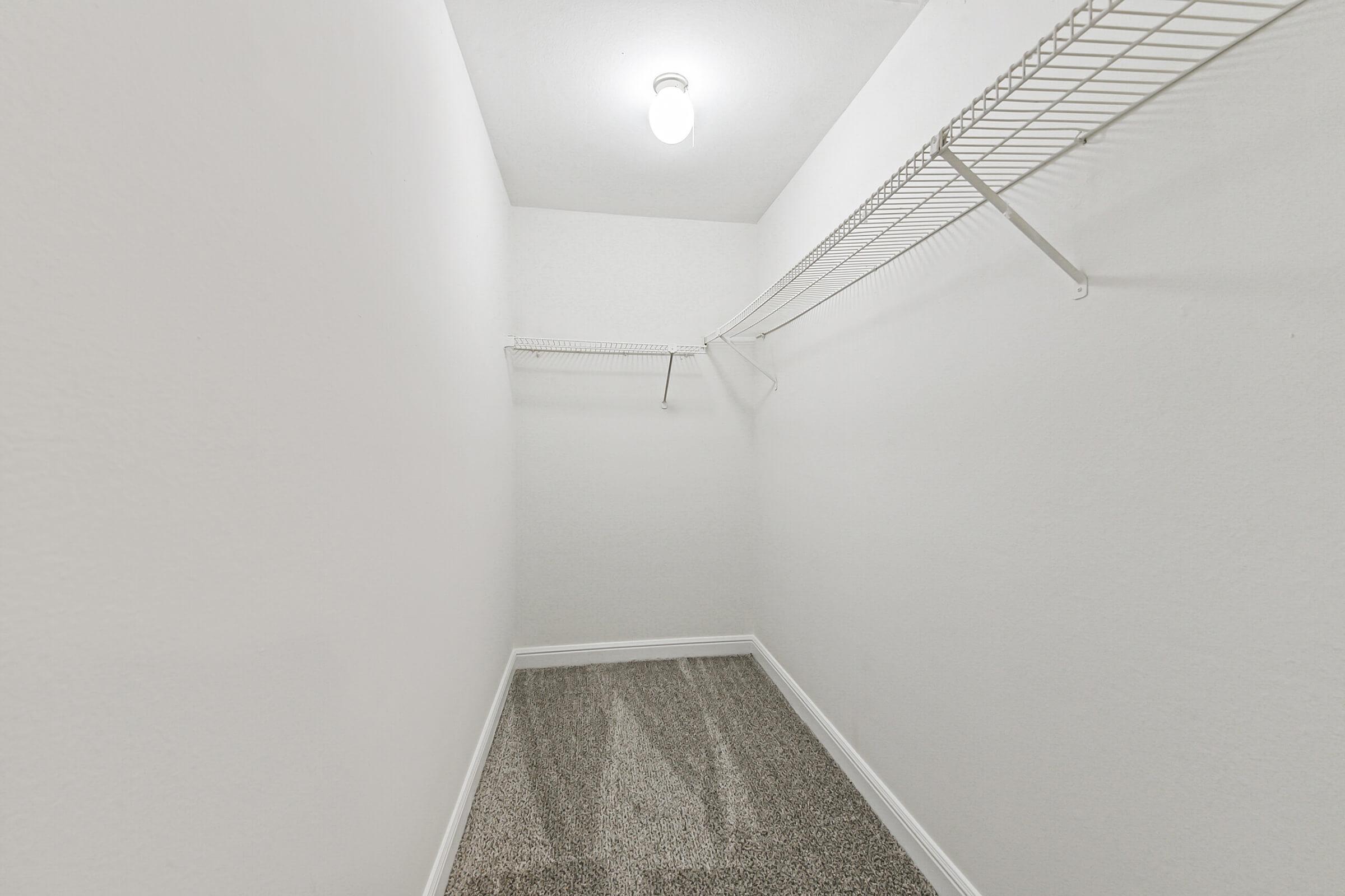 A small, empty walk-in closet with plain white walls and a light bulb overhead. It features a wire shelf on one side and a carpeted floor, creating a clean and minimalistic space.