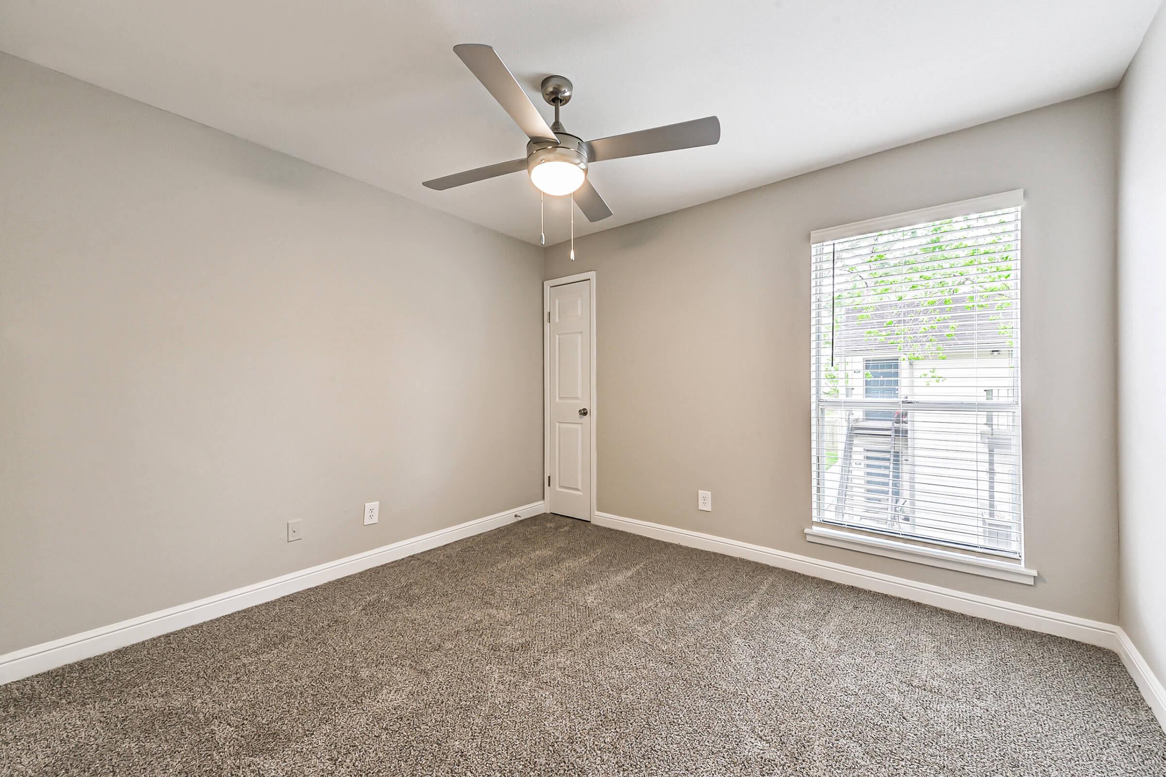 A well-lit, empty bedroom featuring a light gray wall, carpeted floor, and a ceiling fan. There is a closed white door on the left and a window with blinds on the right, allowing natural light to enter. The room has a simple and clean aesthetic, making it suitable for various furnishings.