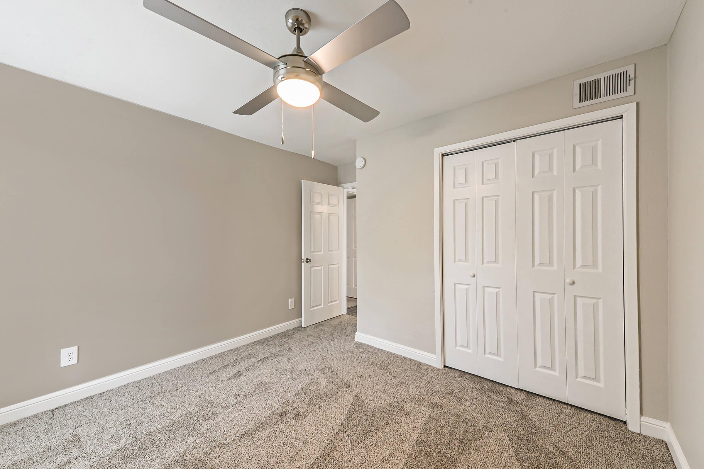 A well-lit room with light gray walls and carpeted floor. The room features a ceiling fan with a light fixture, a partially open door leading to another area, and a closet with double doors. The overall atmosphere is clean and modern, suitable for various uses such as a bedroom or office.