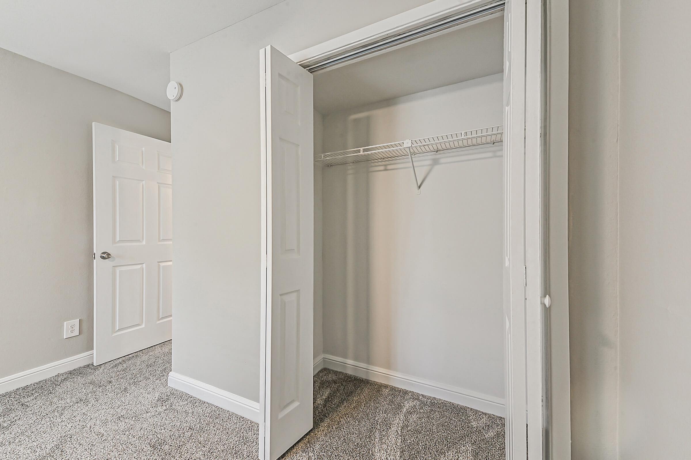 A spacious, empty closet with sliding doors in a well-lit room. The walls are painted a light color, and the floor is covered with grey carpet. A wire shelf is mounted inside the closet, and another door can be seen partially closed in the background.