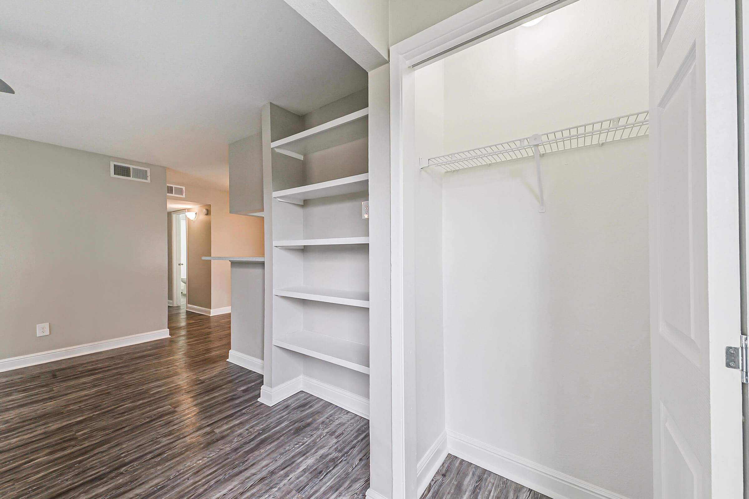 A view of an interior space featuring a closet with open shelving and a hanging rod, light-colored walls, and hardwood-style flooring. The room appears bright and spacious, with a doorway leading to another area in the background.