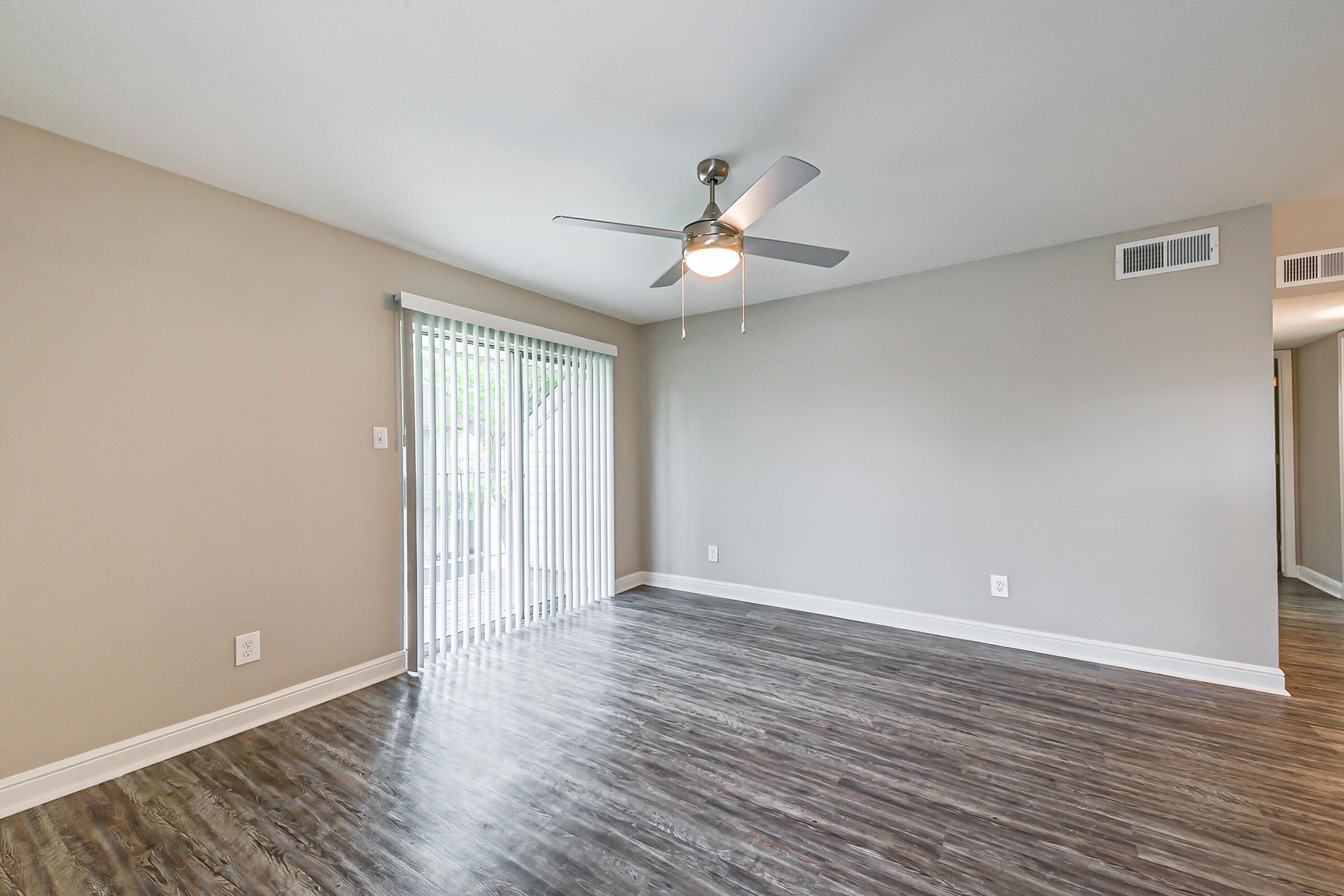 A spacious, empty living room featuring a ceiling fan, light-colored walls, and wood-style flooring. There are large vertical blinds covering a sliding glass door that leads to an outdoor area. The room has a clean, modern aesthetic with natural light coming through the door, creating a bright atmosphere.